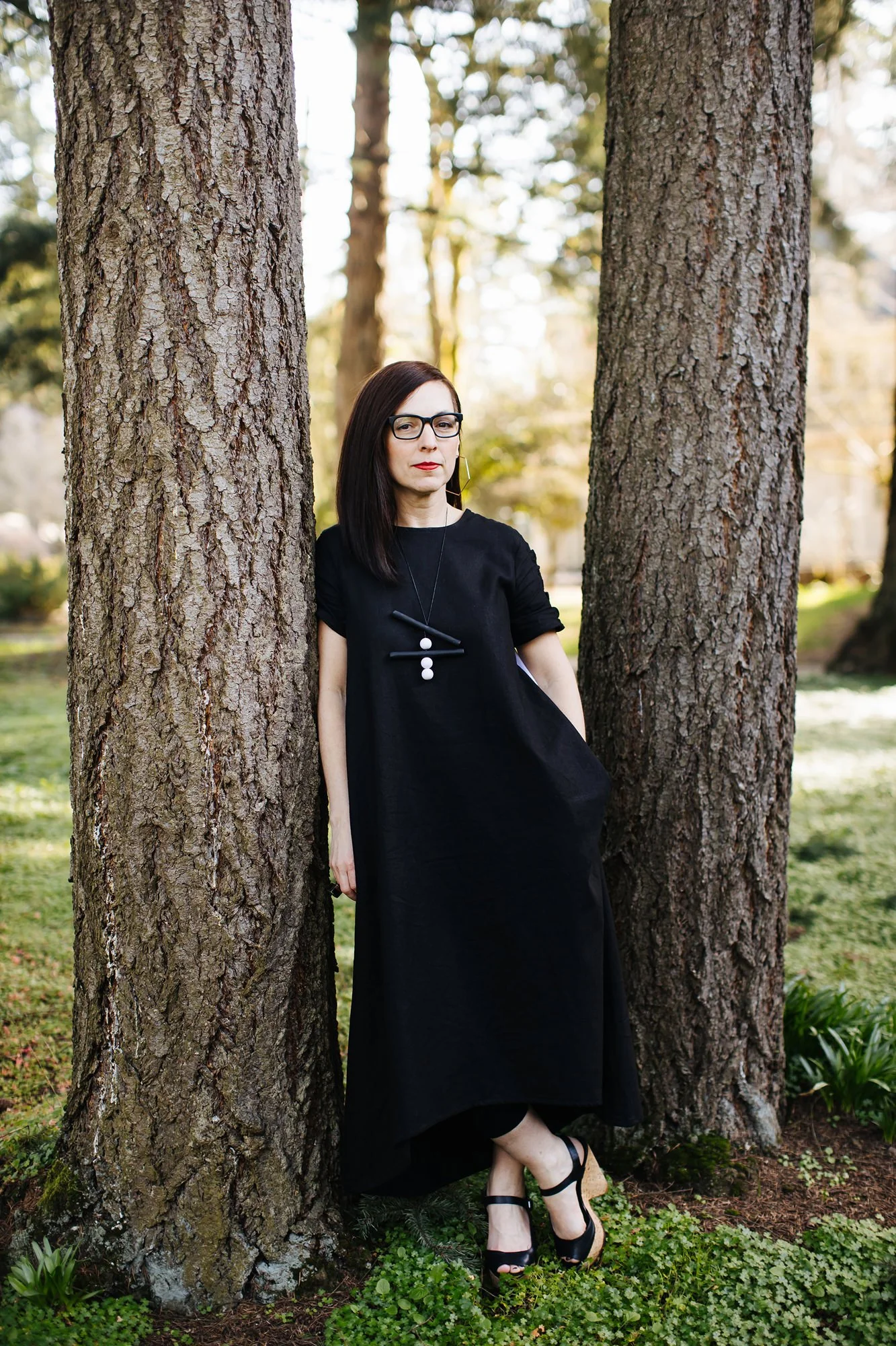 A woman with dark hair and glasses wearing a black dress and heels leaning against a tree in a park.
