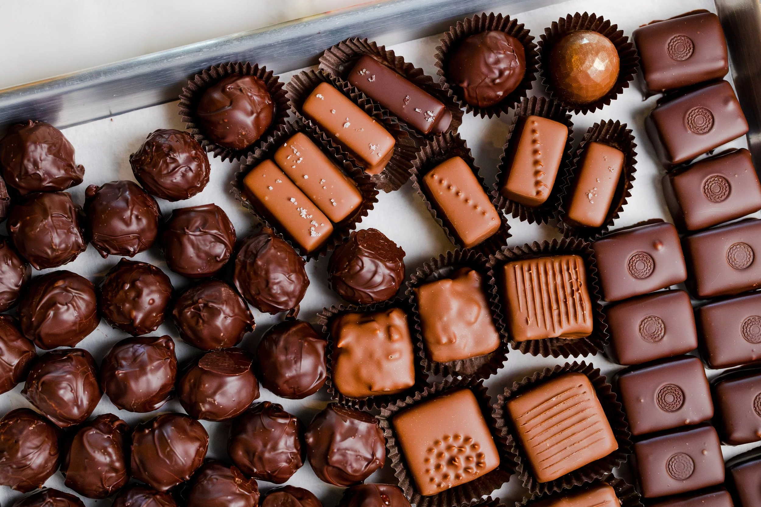 Assorted chocolate candies arranged on a tray, including round truffles, rectangular caramel-filled chocolates, and square chocolates with embossed designs.