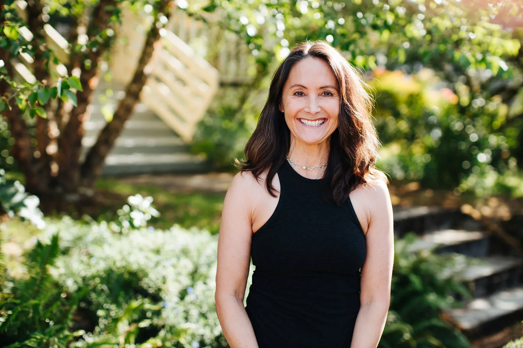 A woman with long brown hair smiling in an outdoor setting with greenery and a staircase in the background.