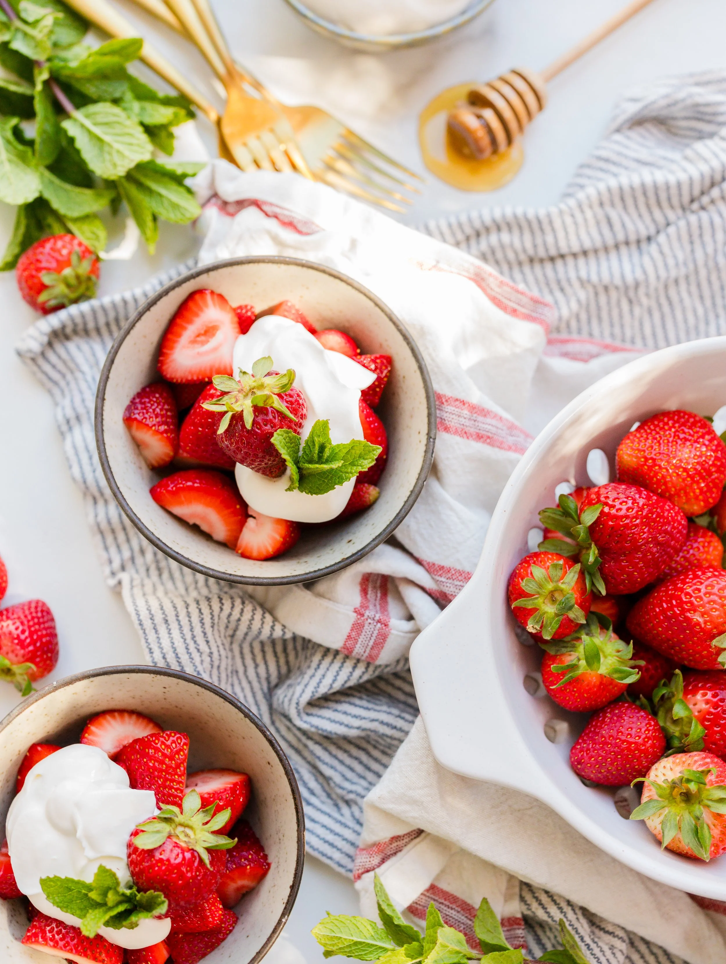Bowl of fresh strawberries topped with whipped cream and mint, surrounded by a bowl of strawberries, honey dipper, and fresh mint leaves on striped cloth and white surface.