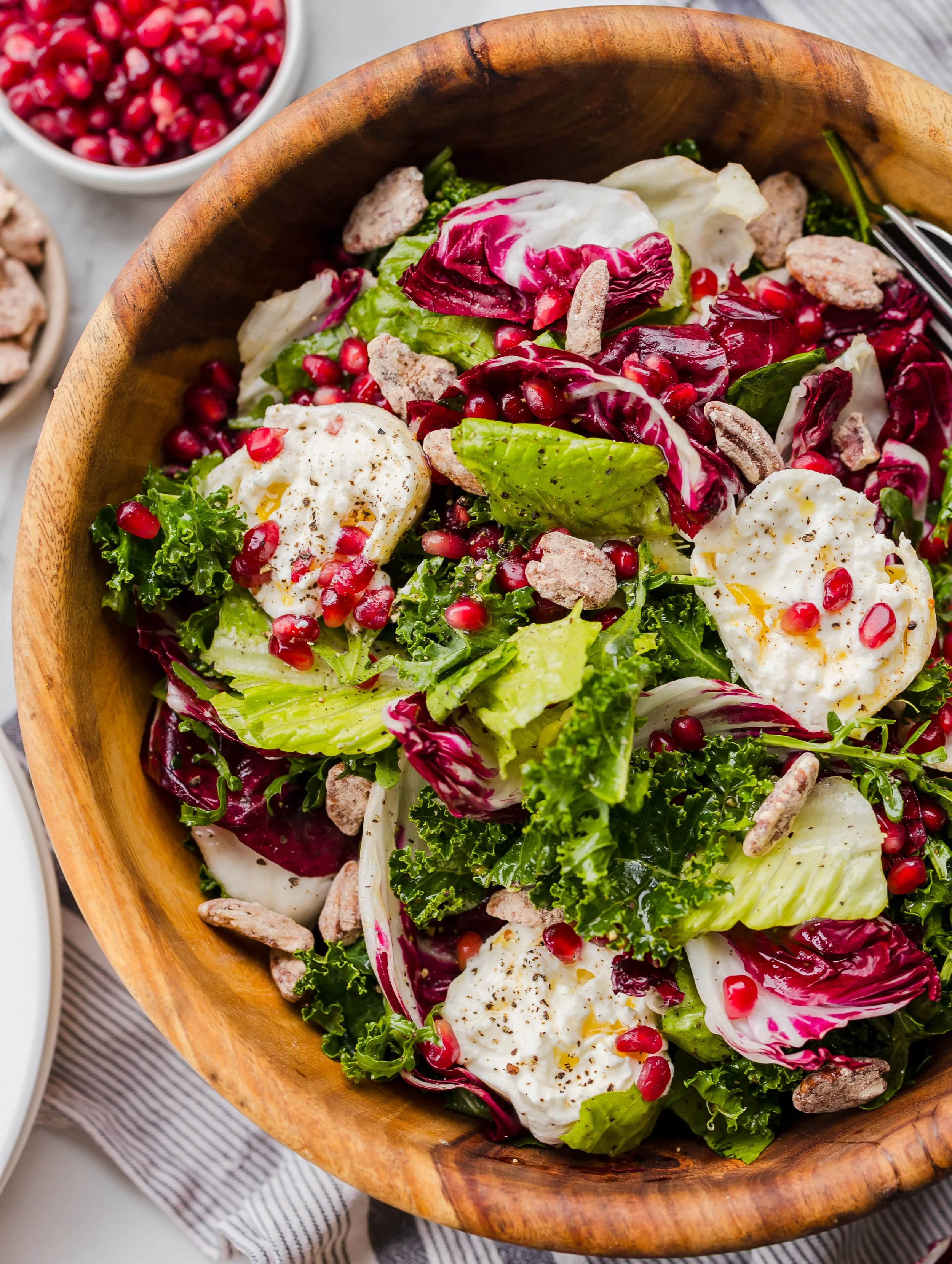 Mixed greens salad with poached eggs, pomegranate seeds, candied pecans, and black pepper in a wooden bowl.