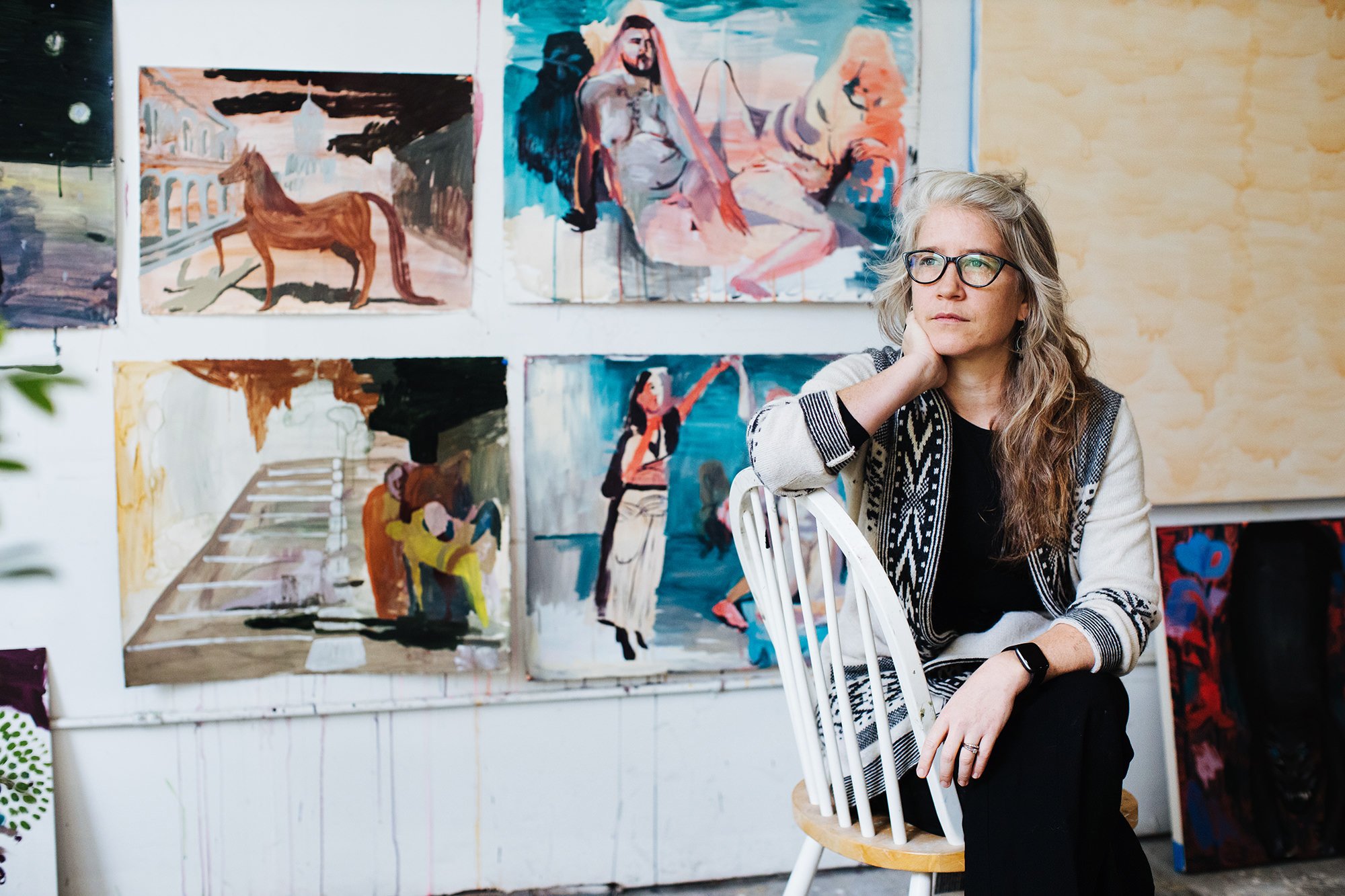 A woman with glasses and long, wavy gray hair sits on a white wooden chair in front of a wall displaying colorful paintings, with her chin resting on her hand, gazing thoughtfully.
