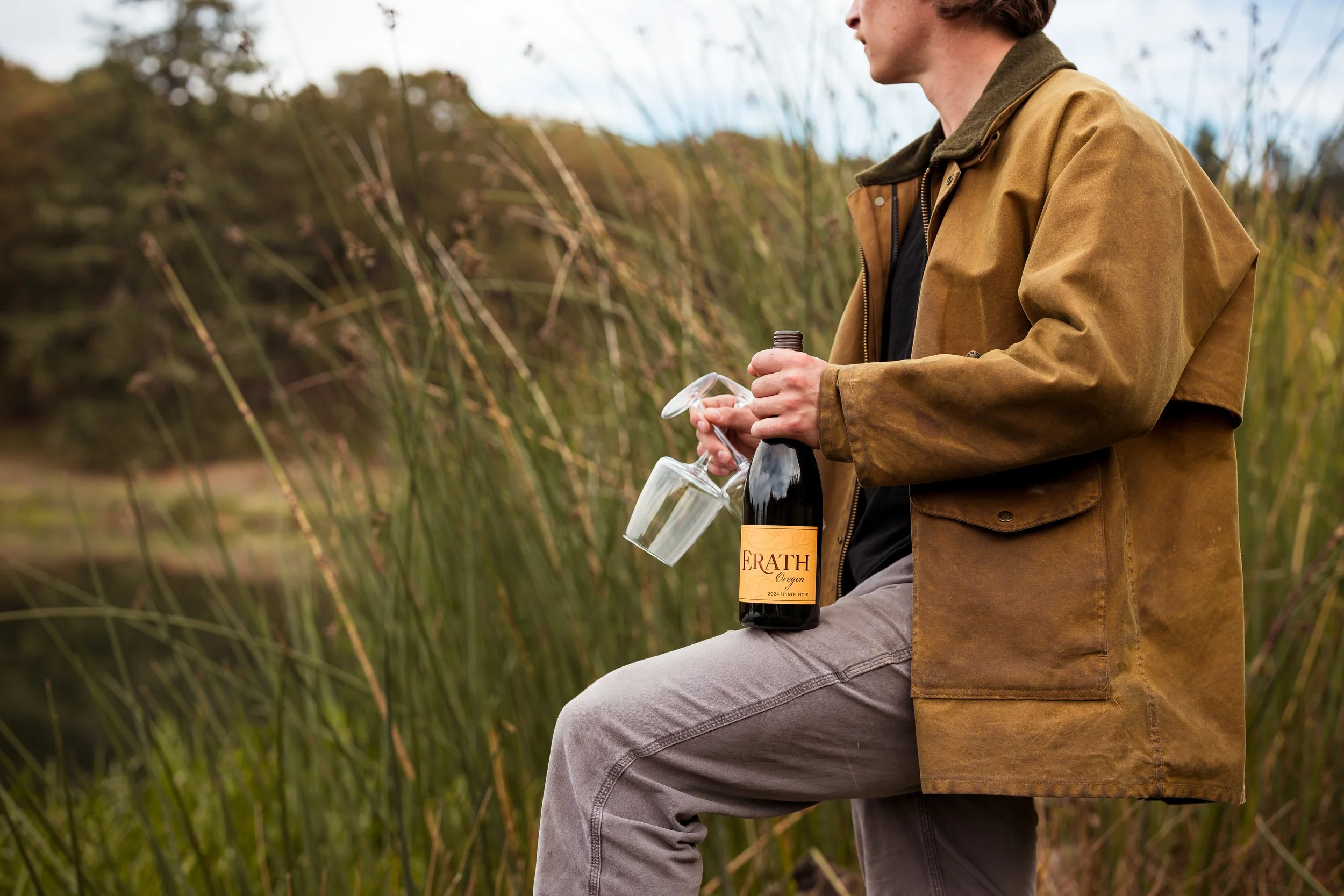 Person holding a wine bottle and glass outdoors in a natural setting with tall grass.