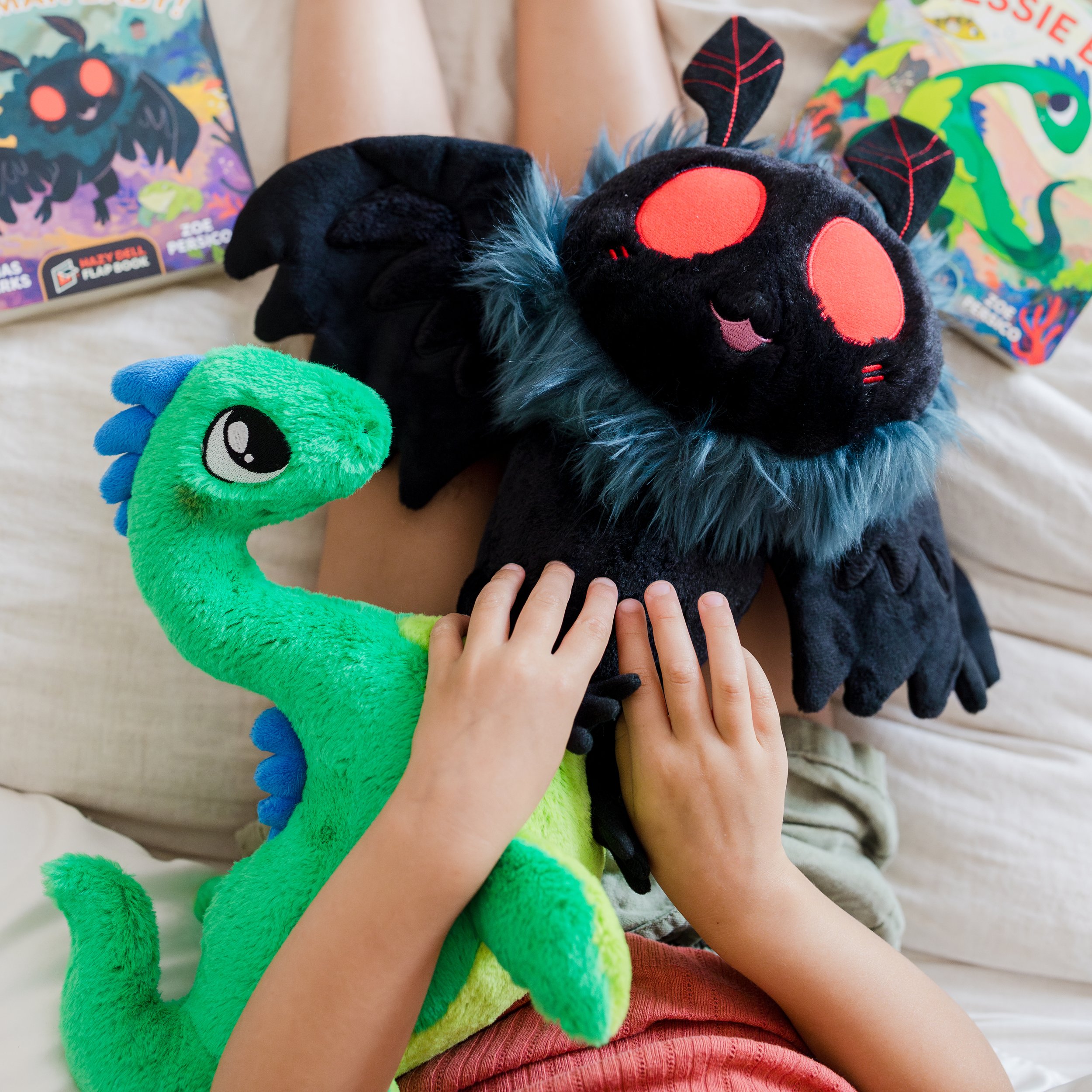 Child sitting on a bed holding plush toys, including a green creature with a big eye and a black plush with red eyes and blue fur details, with colorful books in the background.