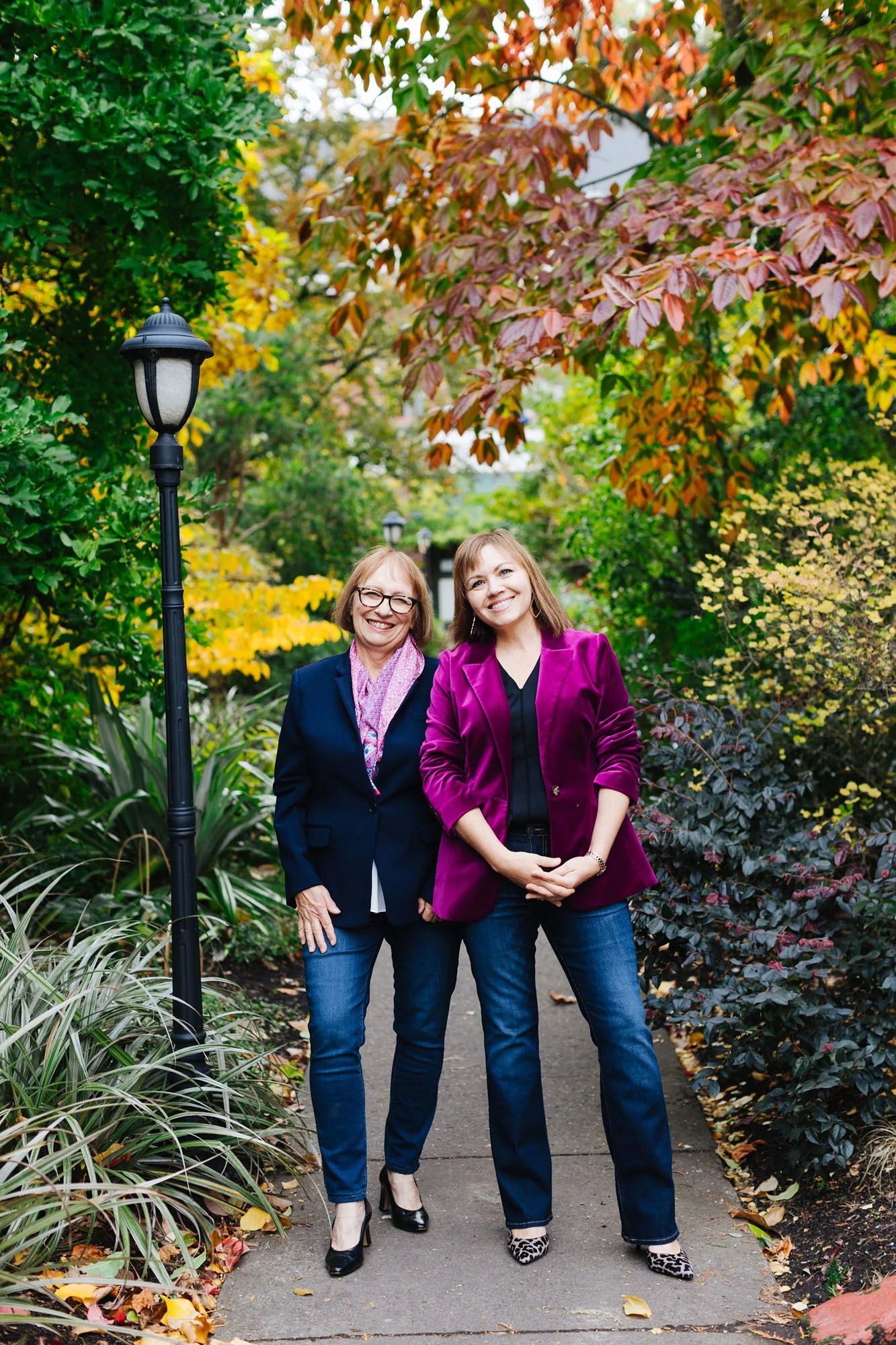 Two women standing on a park path surrounded by vibrant autumn foliage, smiling at the camera. One woman wears a dark blazer, pink scarf, and glasses, and the other wears a magenta blazer and leopard print shoes.