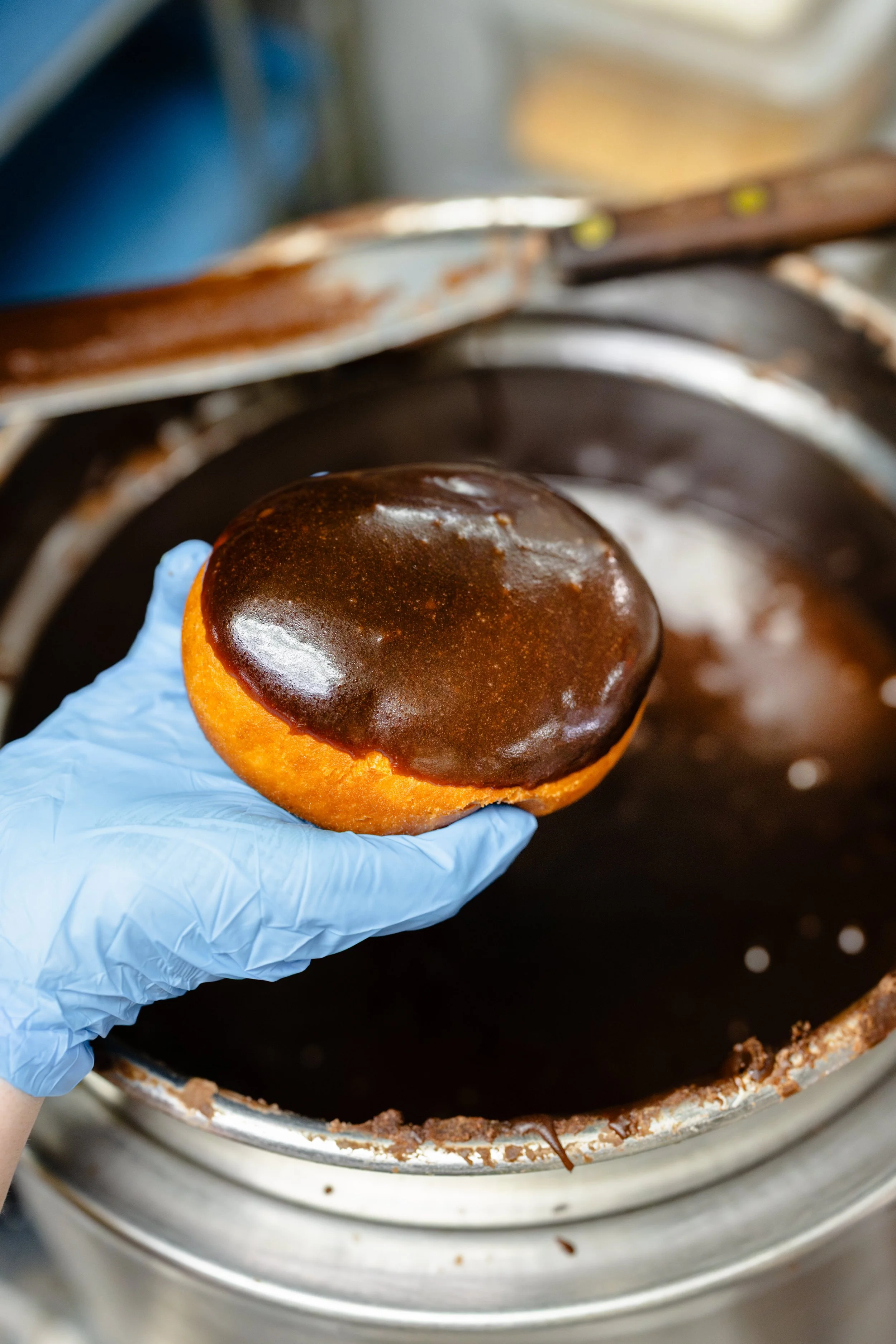 A person wearing a blue glove holds a glazed donut above a chocolate glaze container.