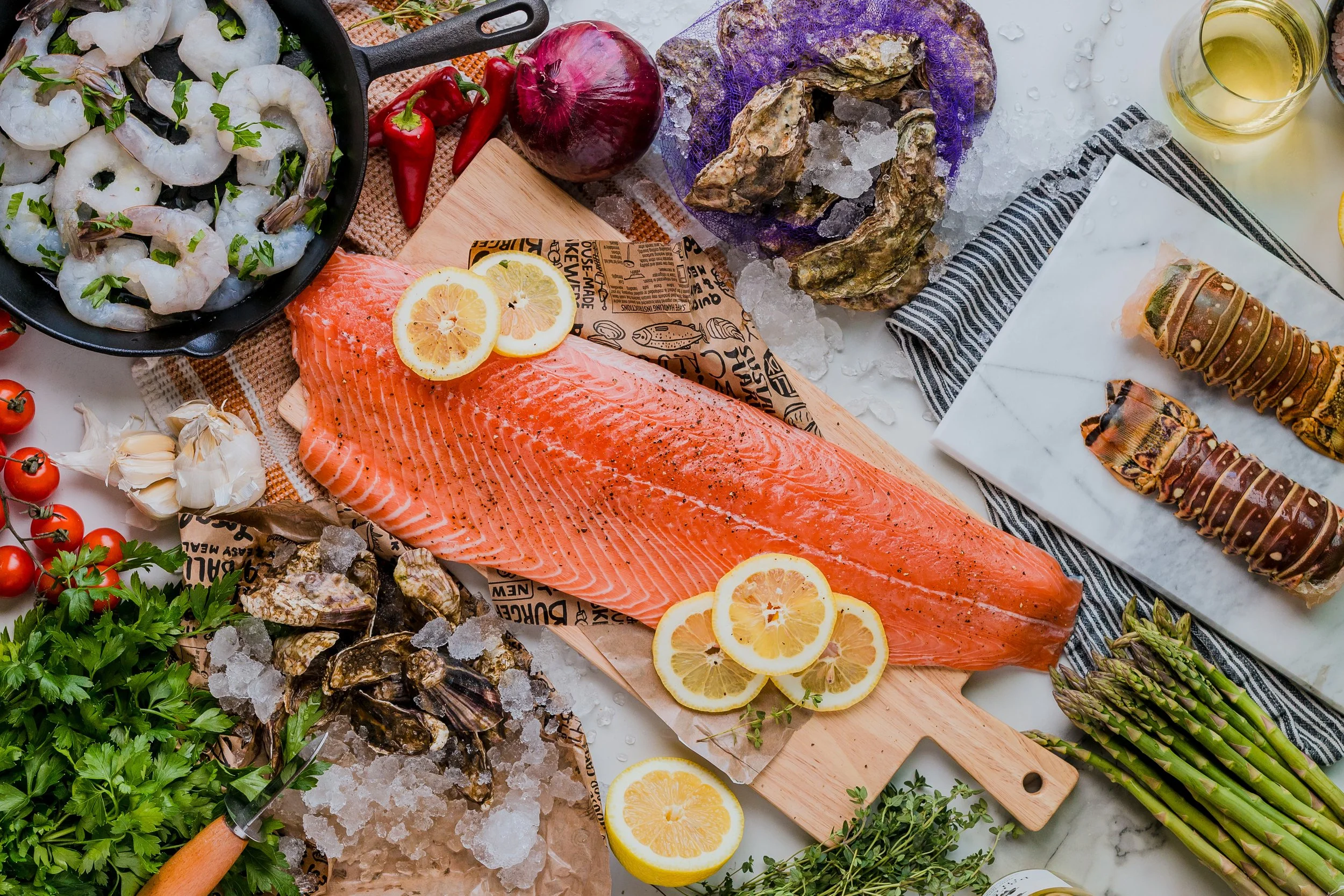 Fresh seafood, vegetables, and herbs on a kitchen countertop, including salmon, shrimp, oysters, lobster, asparagus, cherry tomatoes, garlic, red onion, lemon, and various seasonings.