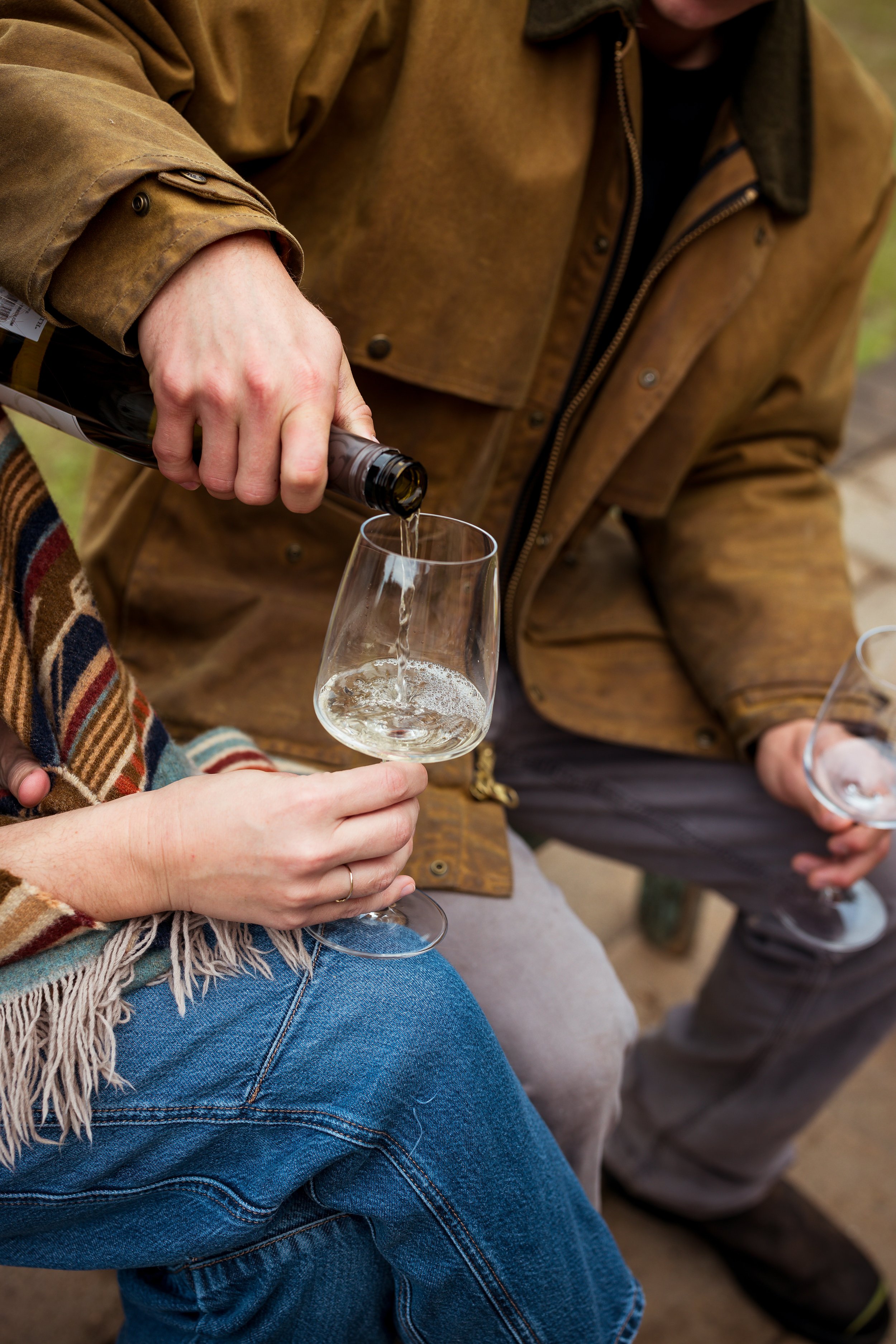 Person pouring champagne into a glass held by another person, both seated outdoors.