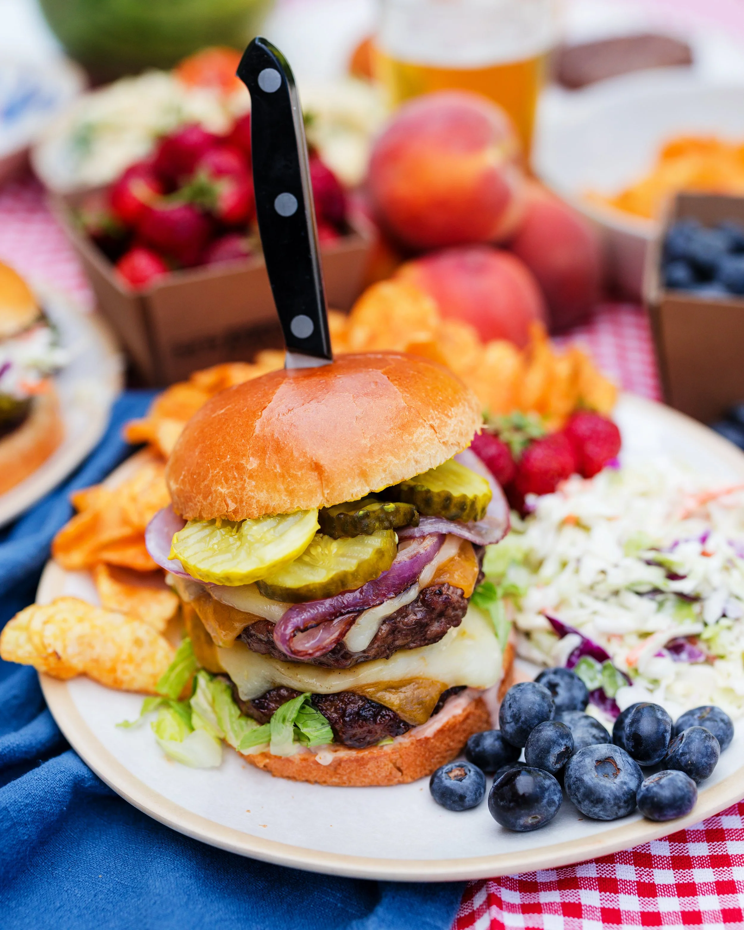A gourmet burger with pickles, onions, lettuce, cheese, and a beef patty in a bun, served on a white plate with blueberries and coleslaw on the side, with fresh fruits and a drink in the background.