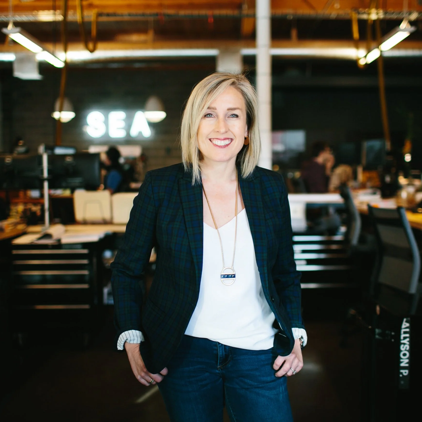 A woman with blonde hair smiling in a modern office or coworking space with the word 'SEA' illuminated on the wall behind her.