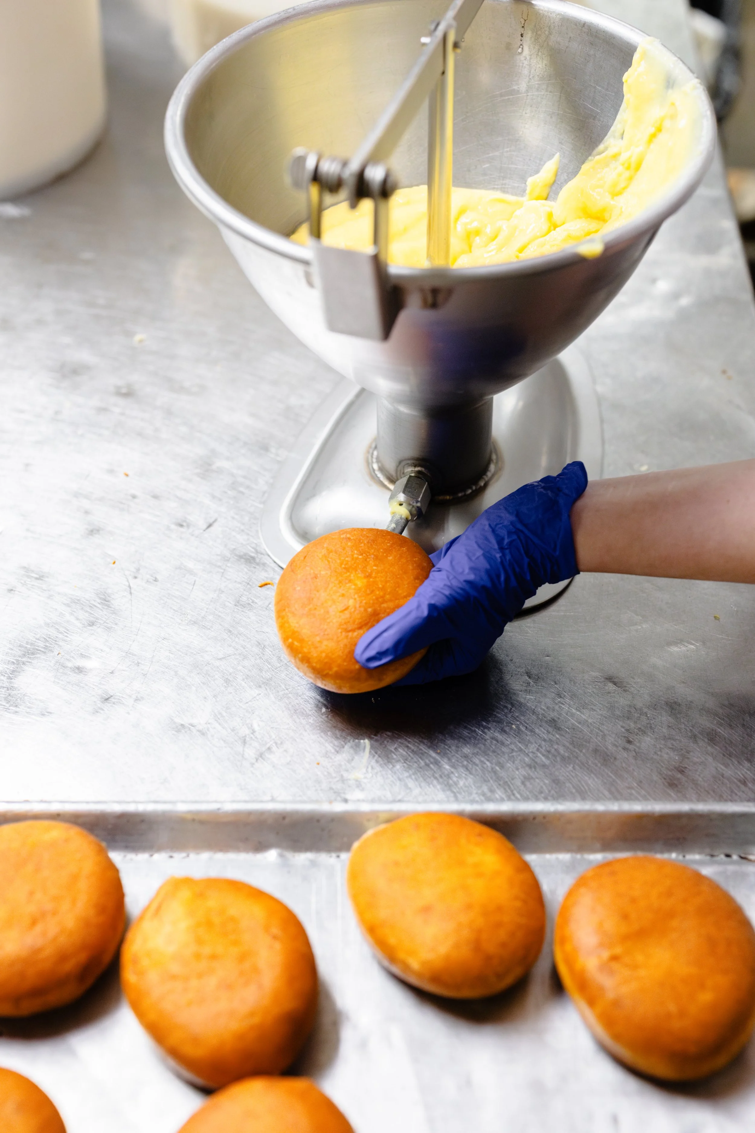 A person wearing a blue glove is filling a round, orange sponge cake with yellow custard or cream using a piping device. There are several similar cakes on a metal tray in the foreground, and a large mixing bowl with more yellow custard or cream in t