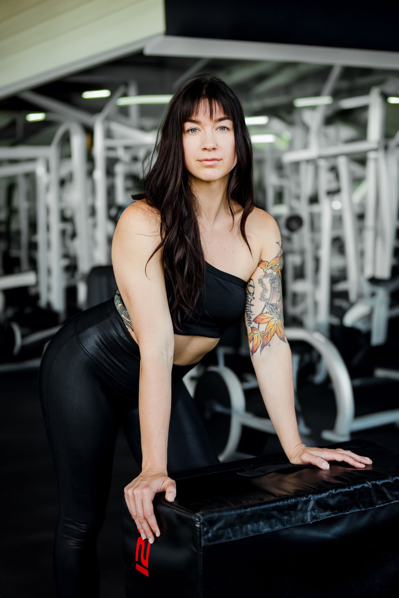 A young woman with long dark hair, tattoos on her left arm, wearing a black sports bra and black leggings, leaning on a black padded workout block in a gym with weight machines in the background.