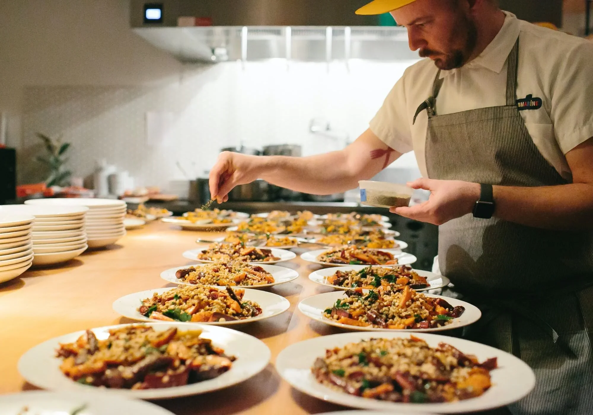 Chef wearing a beige apron and watch, garnishing multiple plated dishes with a sprinkle of herbs or seasoning in a professional kitchen.