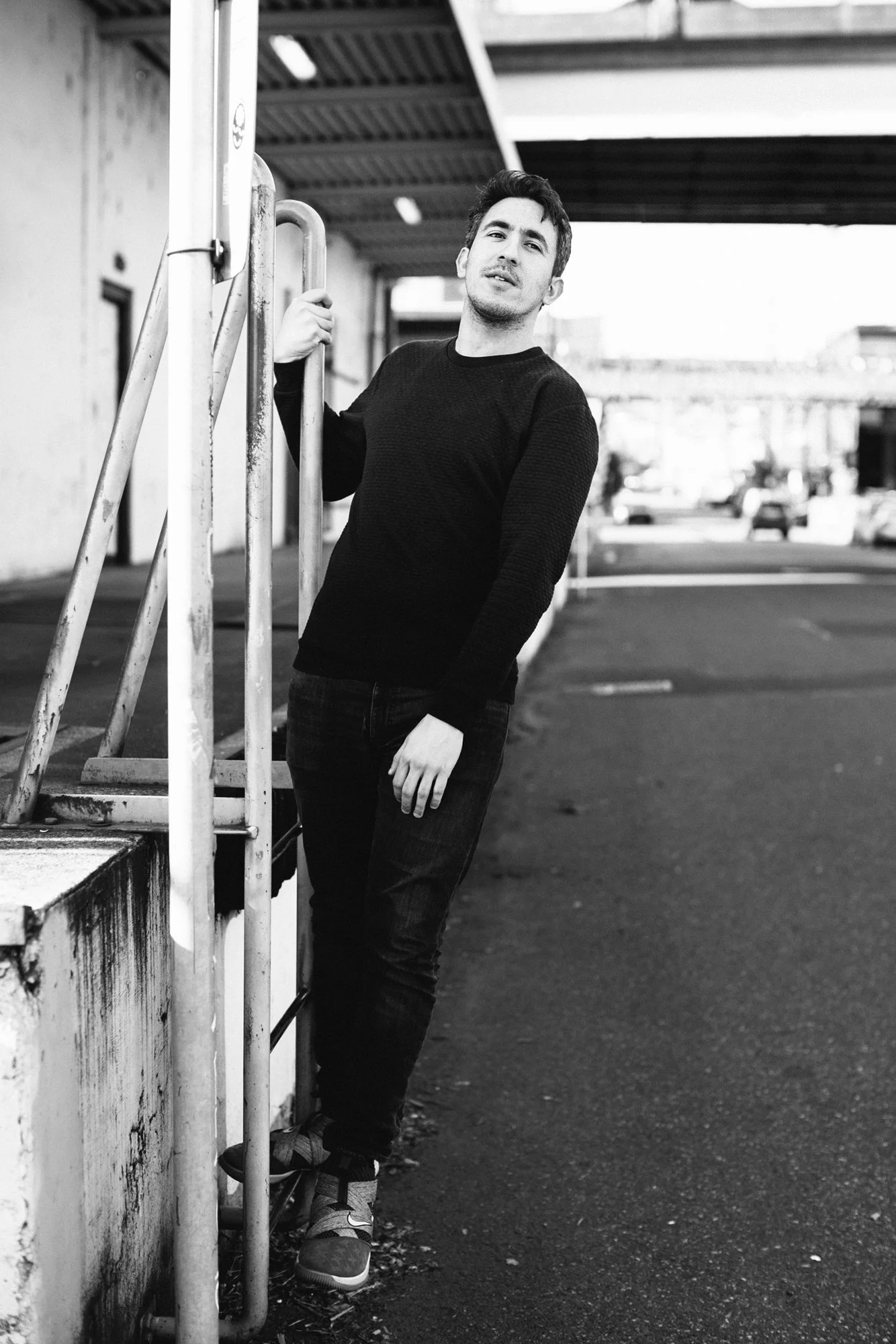 A young man with dark hair, wearing a black sweater, jeans, and sneakers, leaning against a metal railing in an urban setting with cars and buildings in the background, in black and white.