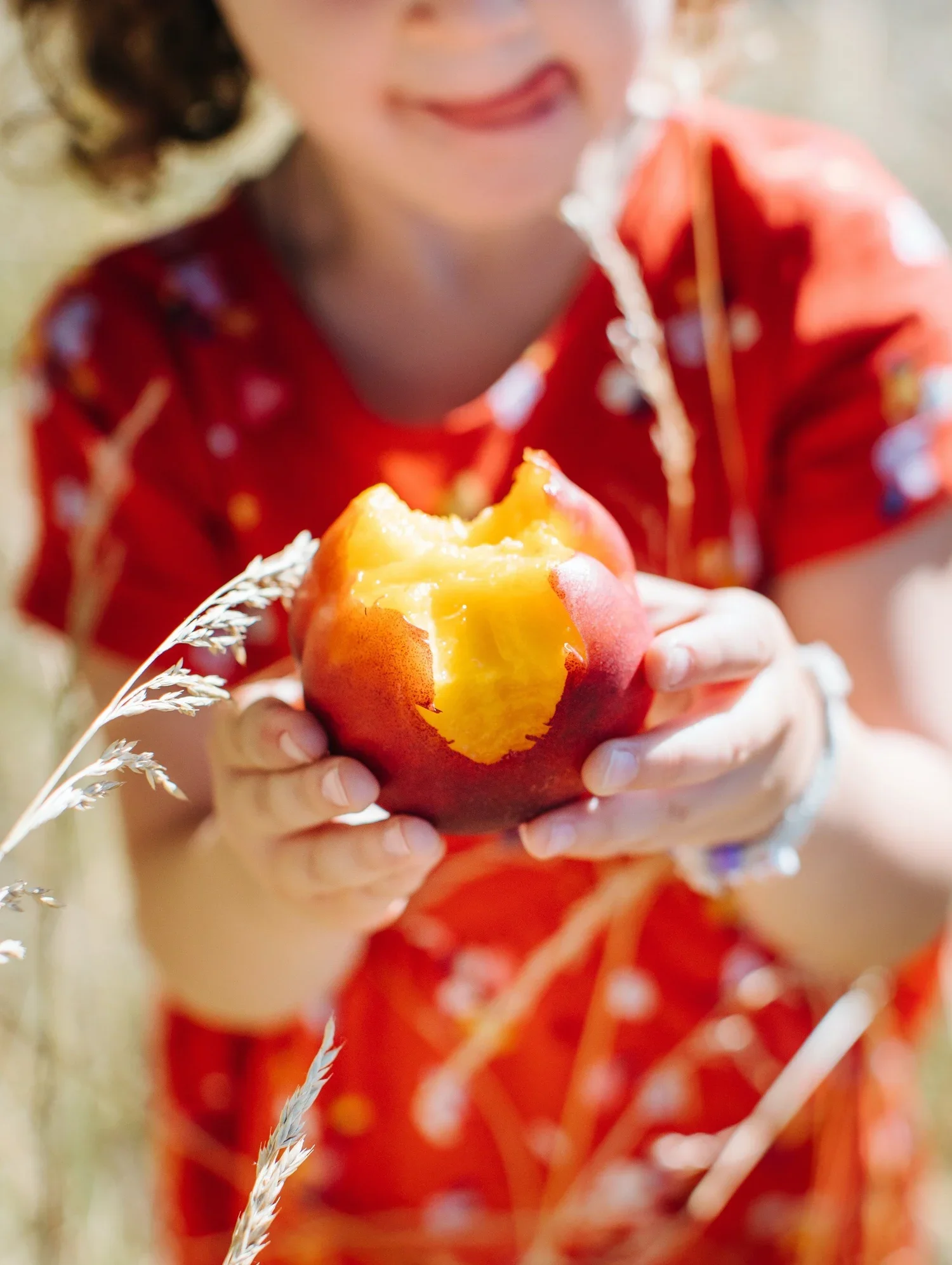 A person in an orange dress with white patterns holds a bitten peach in front of them, with a blurred background.