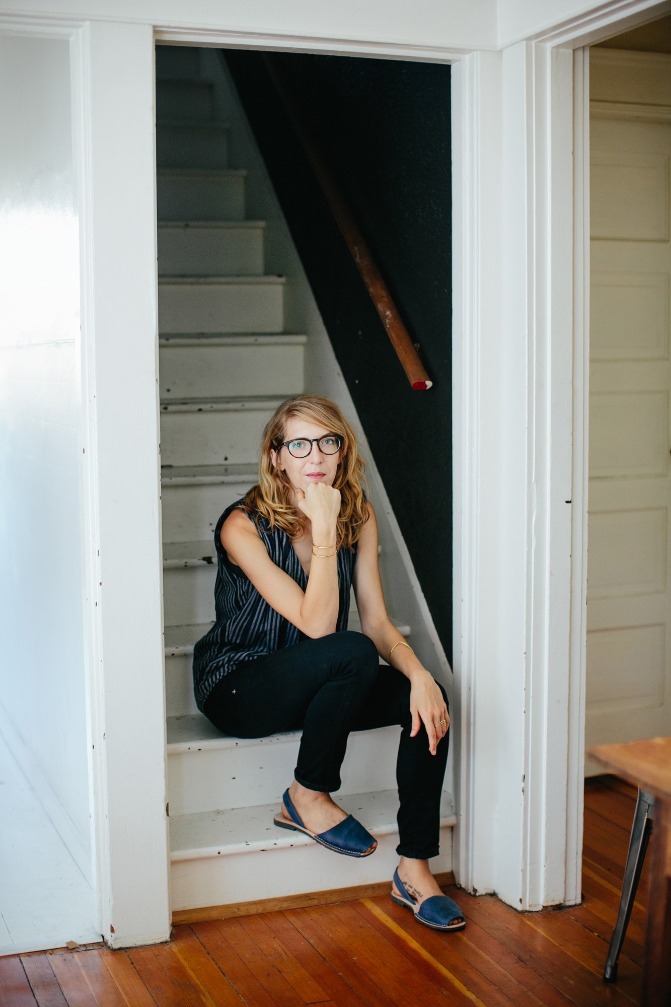 A woman with glasses and blonde hair, wearing a sleeveless striped top, dark pants, and blue open-toed sandals, sitting on white stairs in a doorway, with a dark wall and a wooden stair stick behind her.