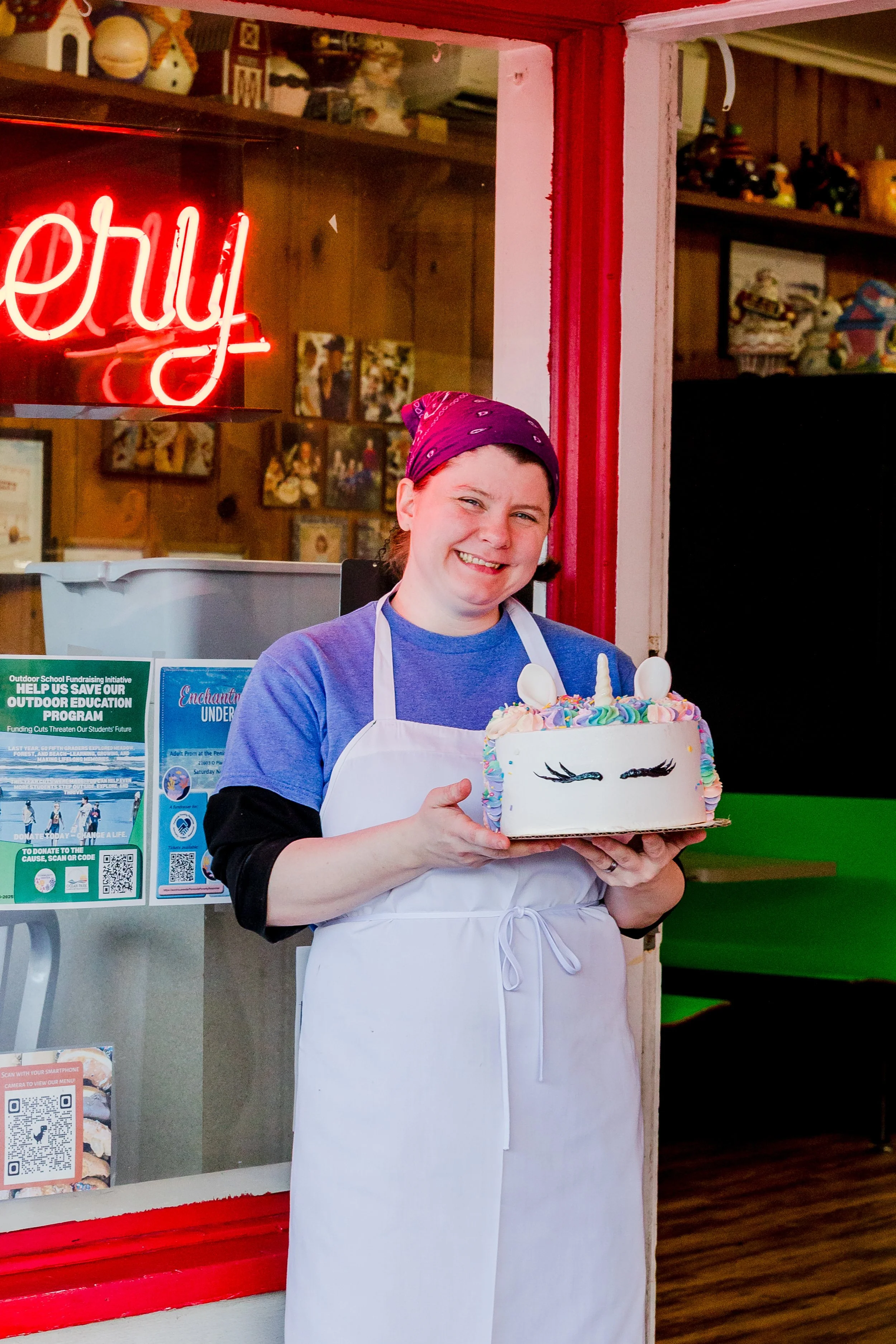 A person holding a unicorn-themed birthday cake with a smiling face, rainbow-colored decorations, and unicorn horns, standing inside a bakery or cafe.