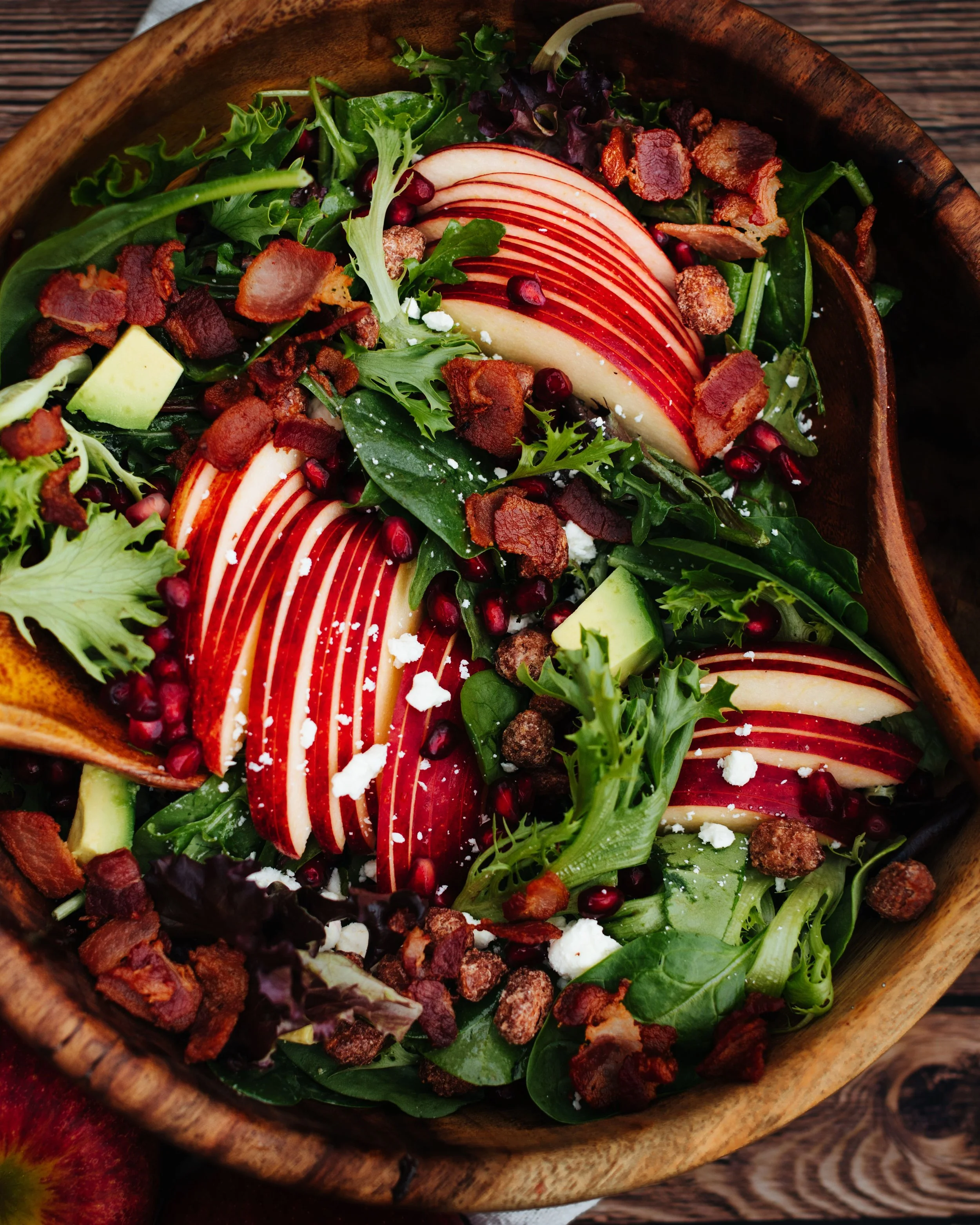 Fresh mixed salad with sliced apples, bacon bits, pomegranate seeds, leafy greens, and crumbled cheese in a wooden bowl.