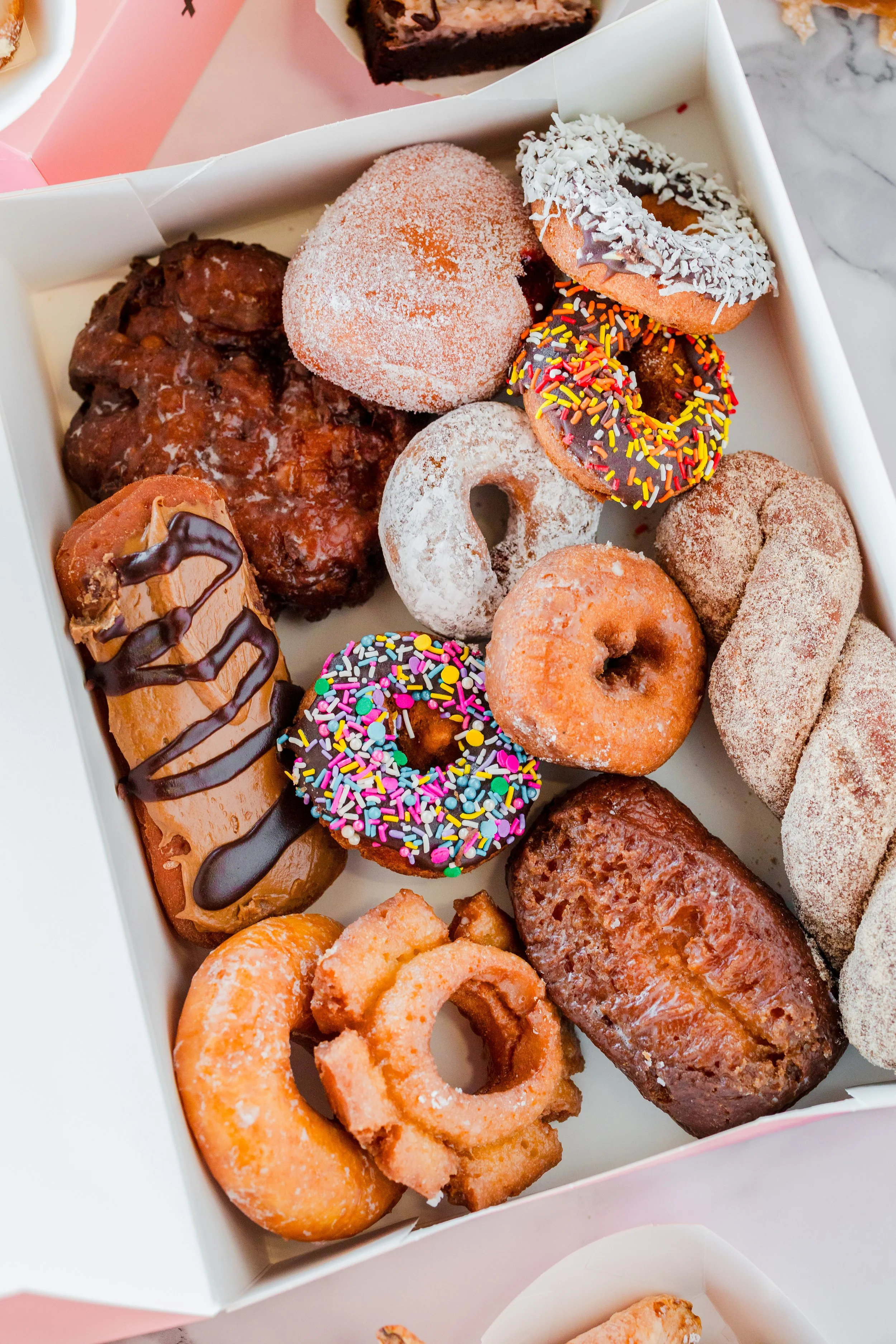 Assorted donuts with various toppings in a box, including sprinkles, chocolate drizzle, powdered sugar, and cinnamon sugar.