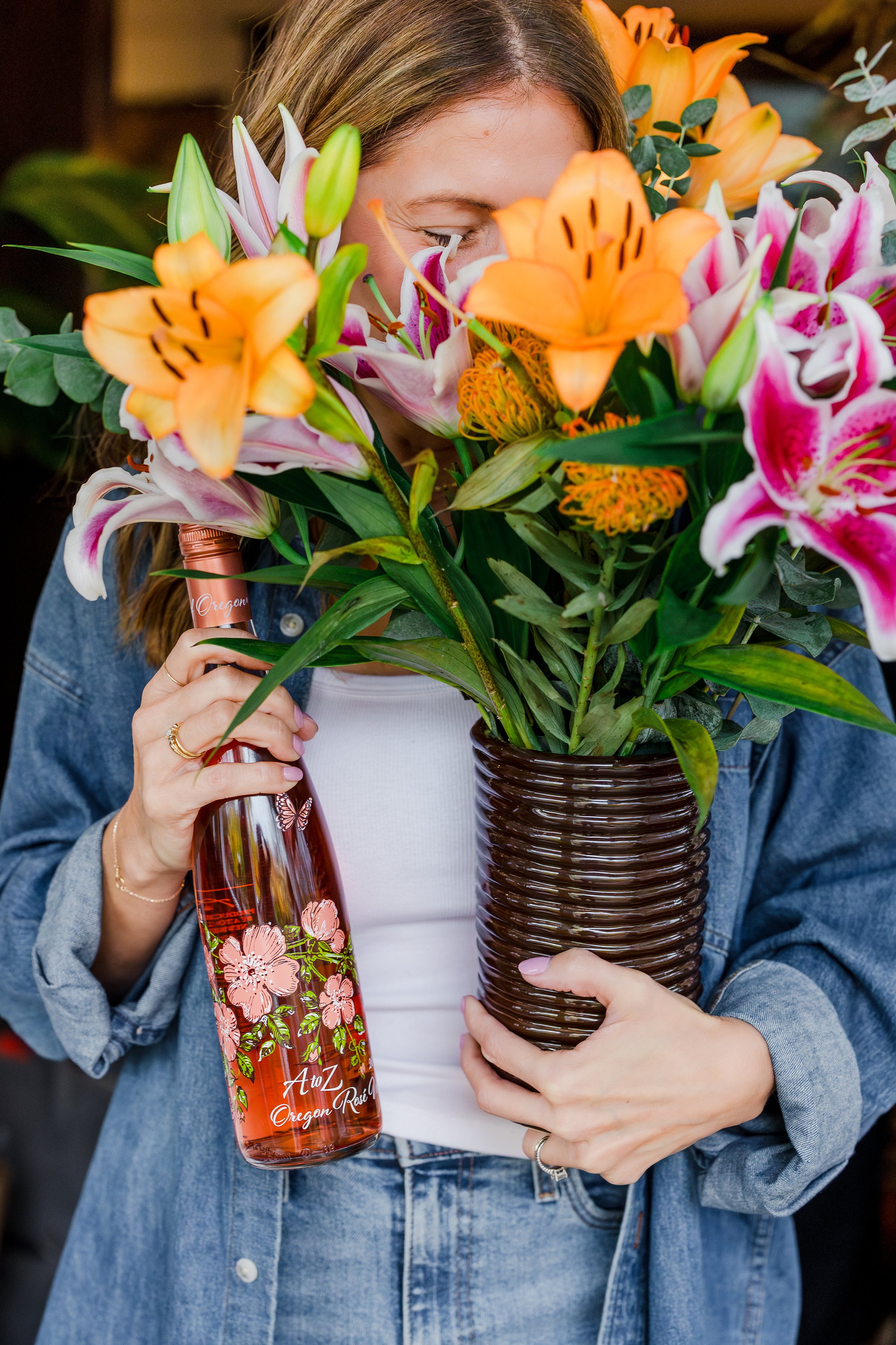 Person holding a large bouquet of vibrant pink, orange, and white lilies in a brown woven vase and a bottle of rosé wine decorated with pink flowers and the text 'AutZ Oregon Rose'.