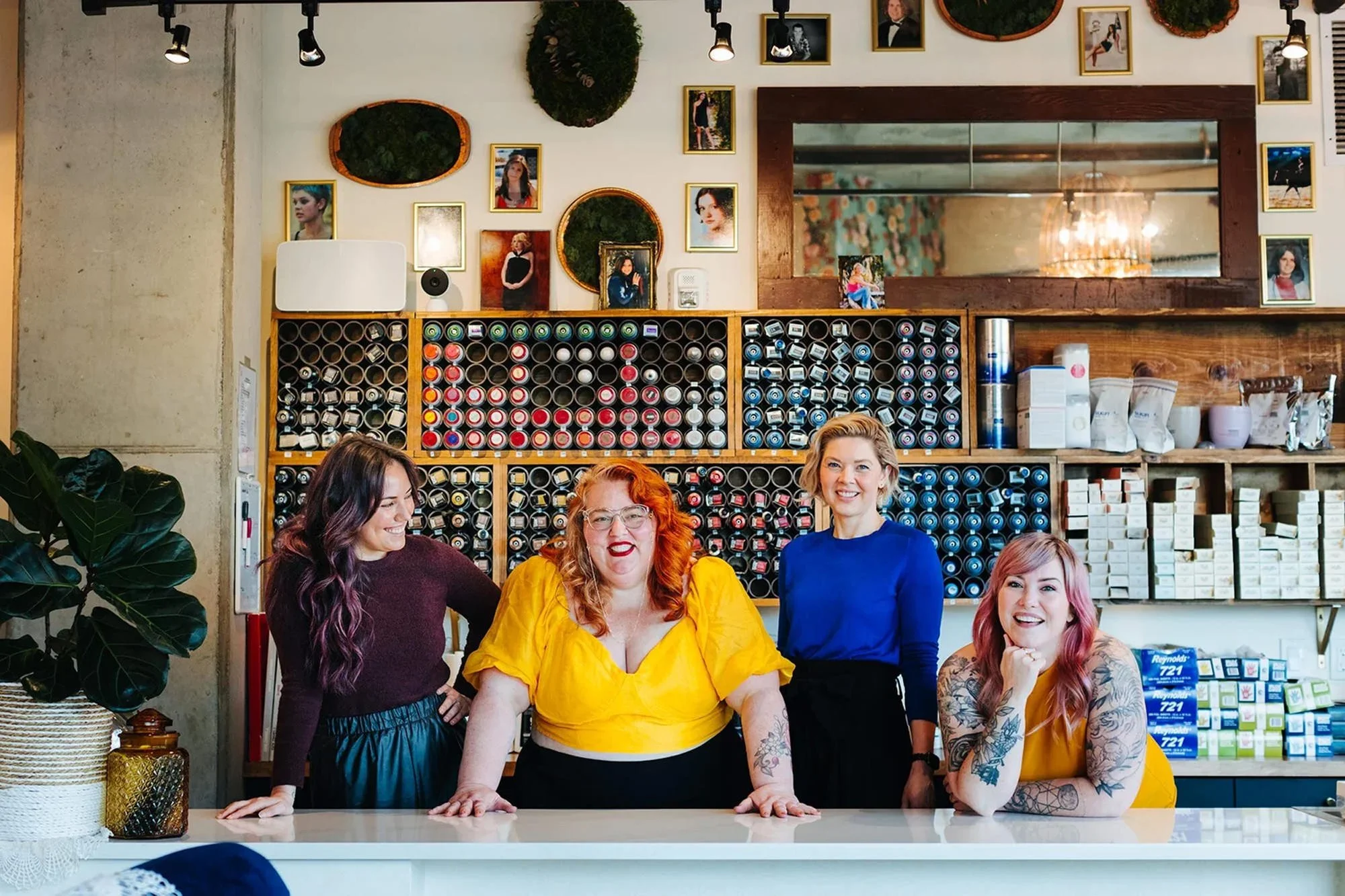 Four women standing behind a counter in a boutique or salon, smiling, with shelves of hair products and wall photos behind them.