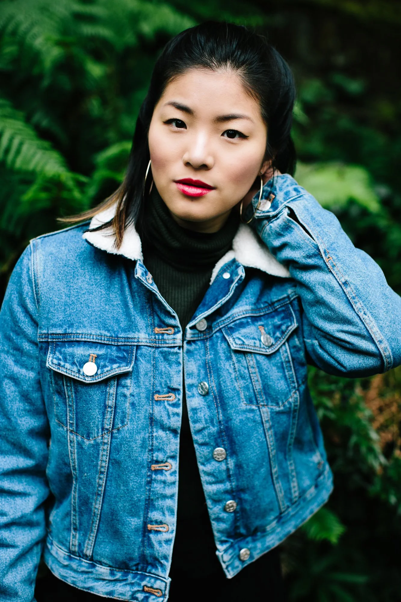 A woman with dark hair wearing a denim jacket with a furry collar, a black turtleneck, earrings, and red lipstick, standing outdoors with green foliage in the background.