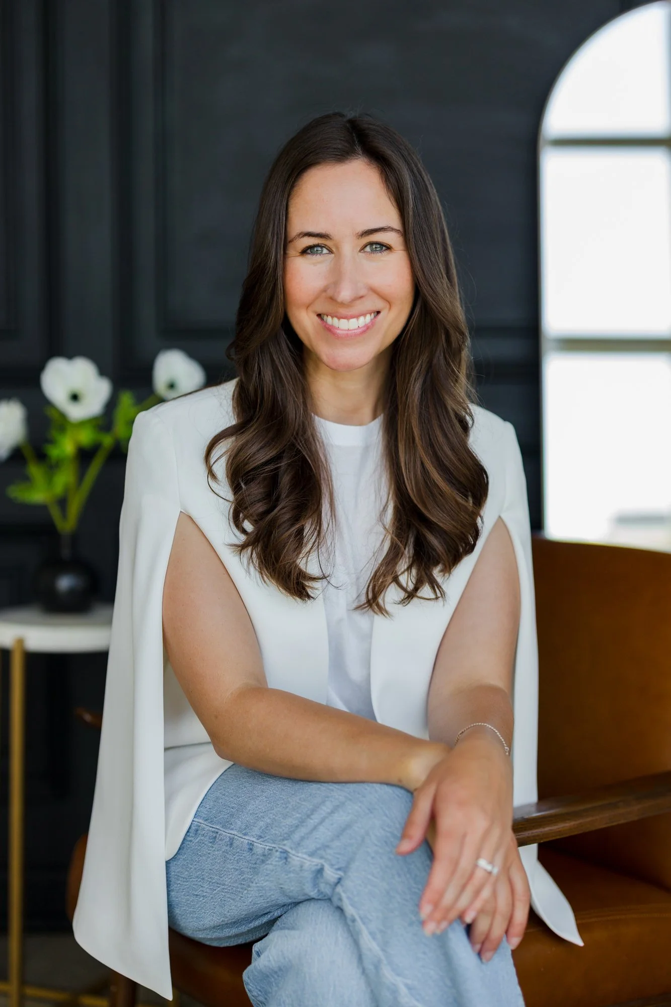 A woman with long wavy brown hair, smiling, sitting indoors on a brown chair with a black wall background and a large window behind her. She is wearing a white sleeveless vest over a white top and light blue jeans.