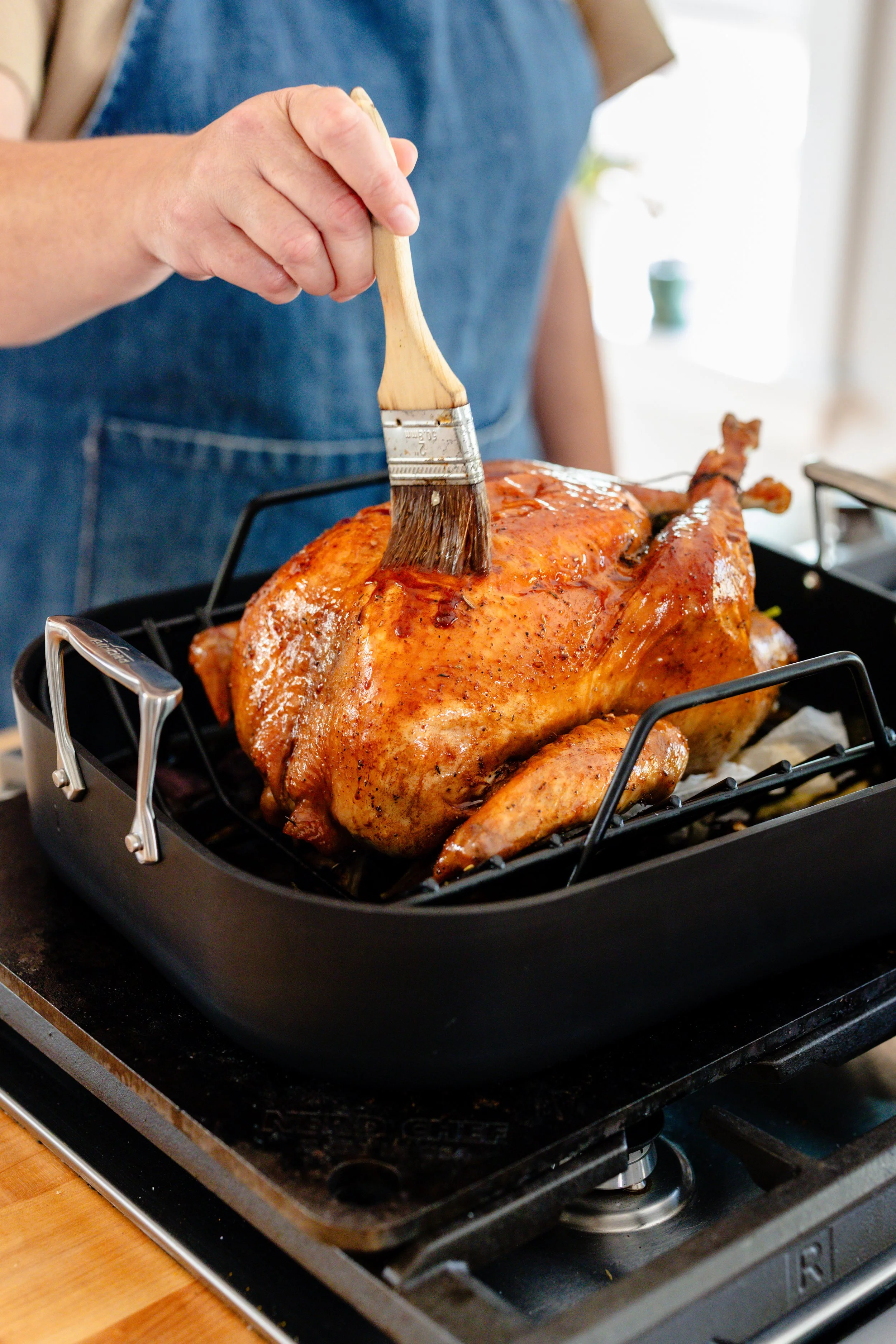 Person brushing glaze or sauce onto a roasted whole chicken in a black roasting pan.
