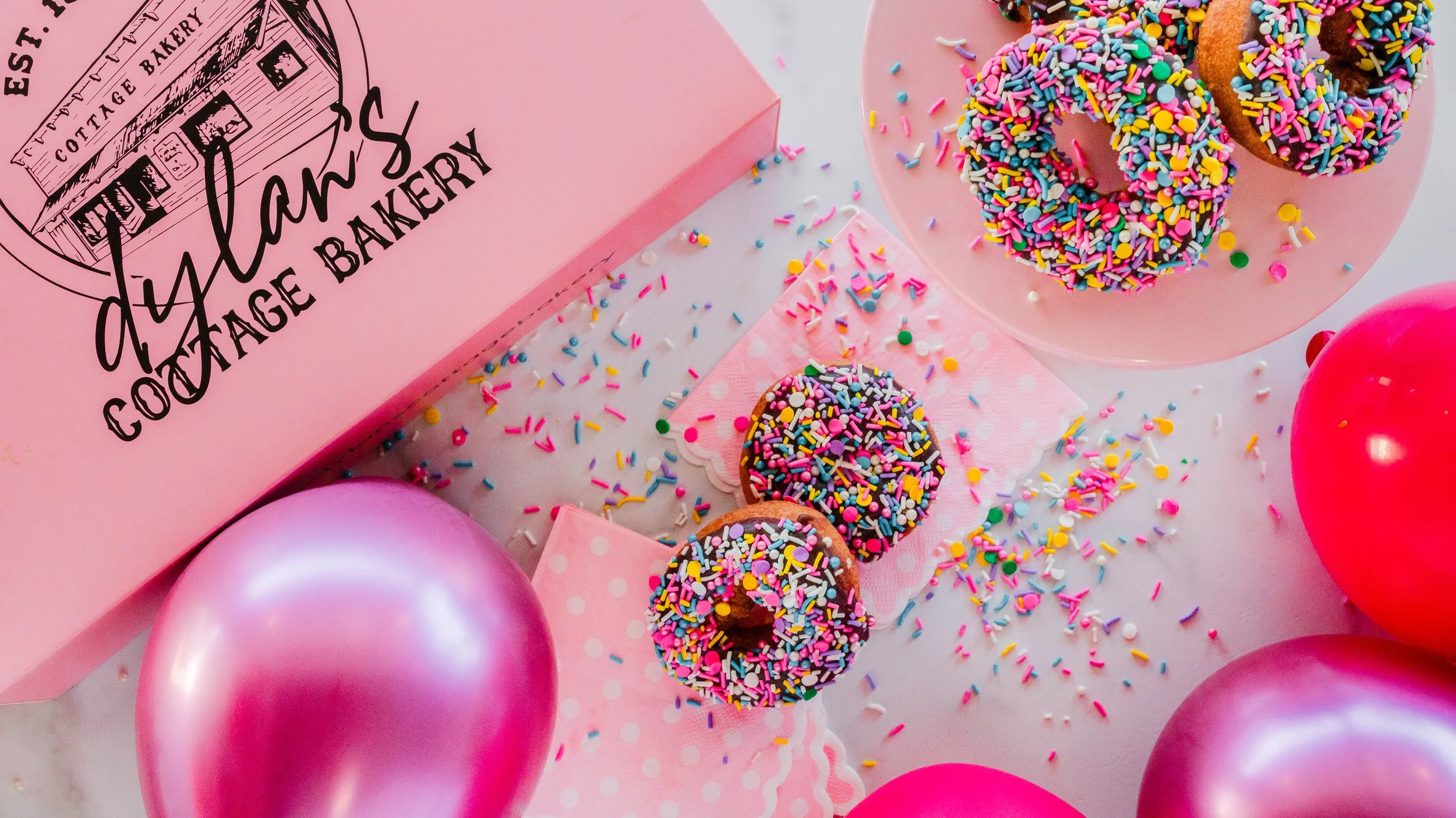Colorful doughnuts with rainbow sprinkles on a pink plate, pink and purple balloons, and a pink bakery box with black lettering that says "Yolanda's Courage Bakery."
