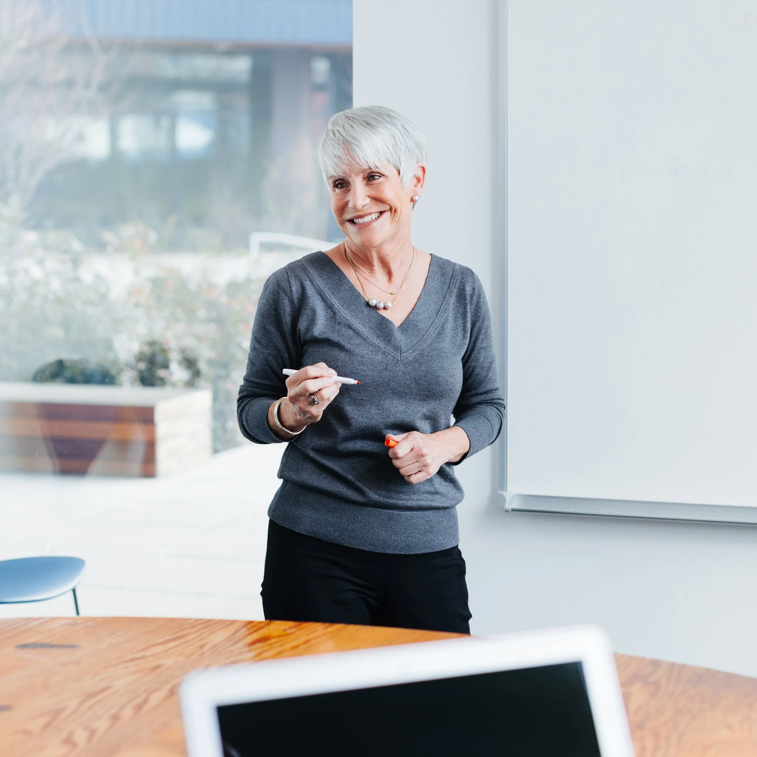 A smiling woman with short gray hair, wearing a gray top and pearl necklace, standing in a bright room near a whiteboard and large window, holding a marker.