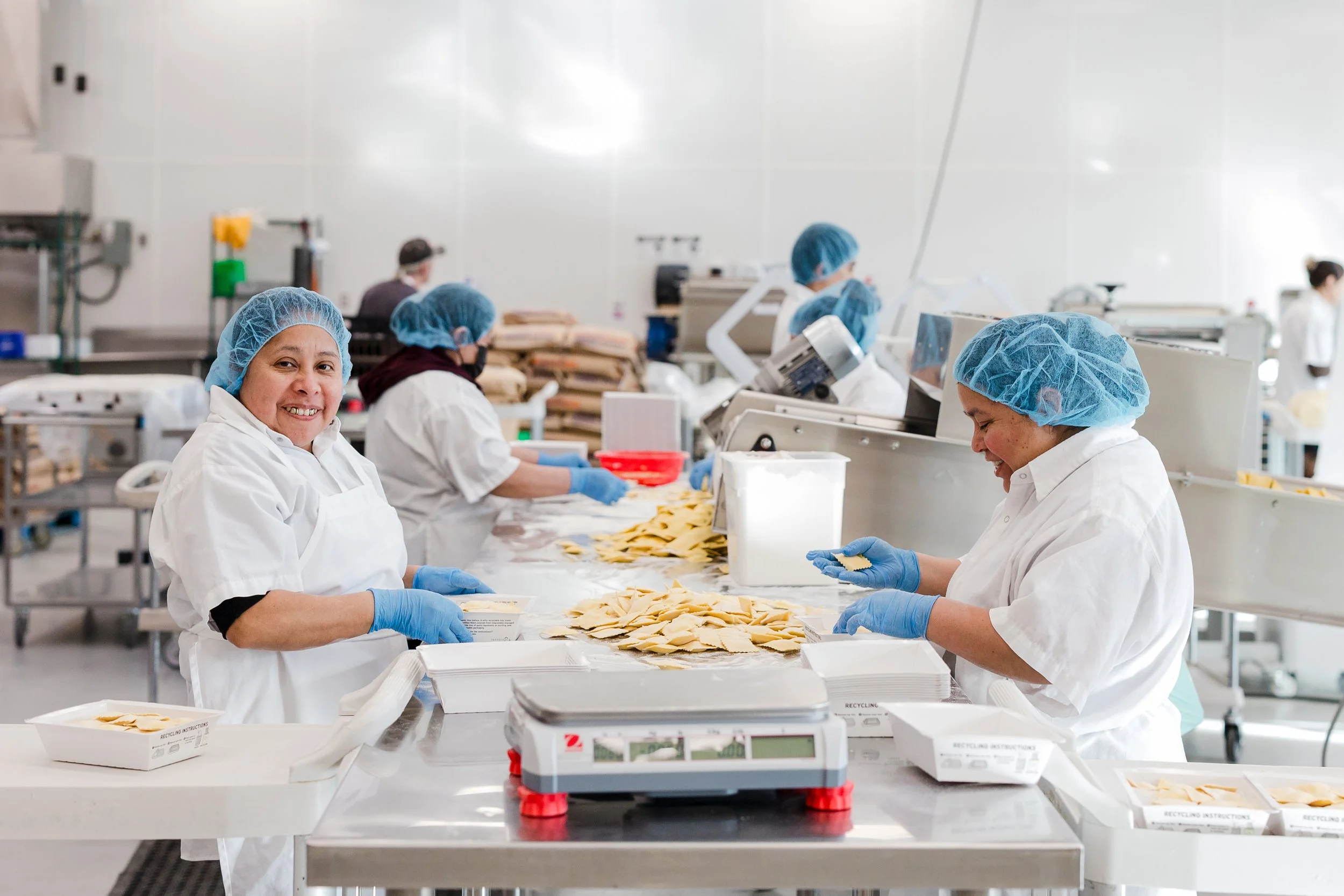 Workers in white uniforms and blue hairnets working on food packaging in a clean, industrial kitchen environment.