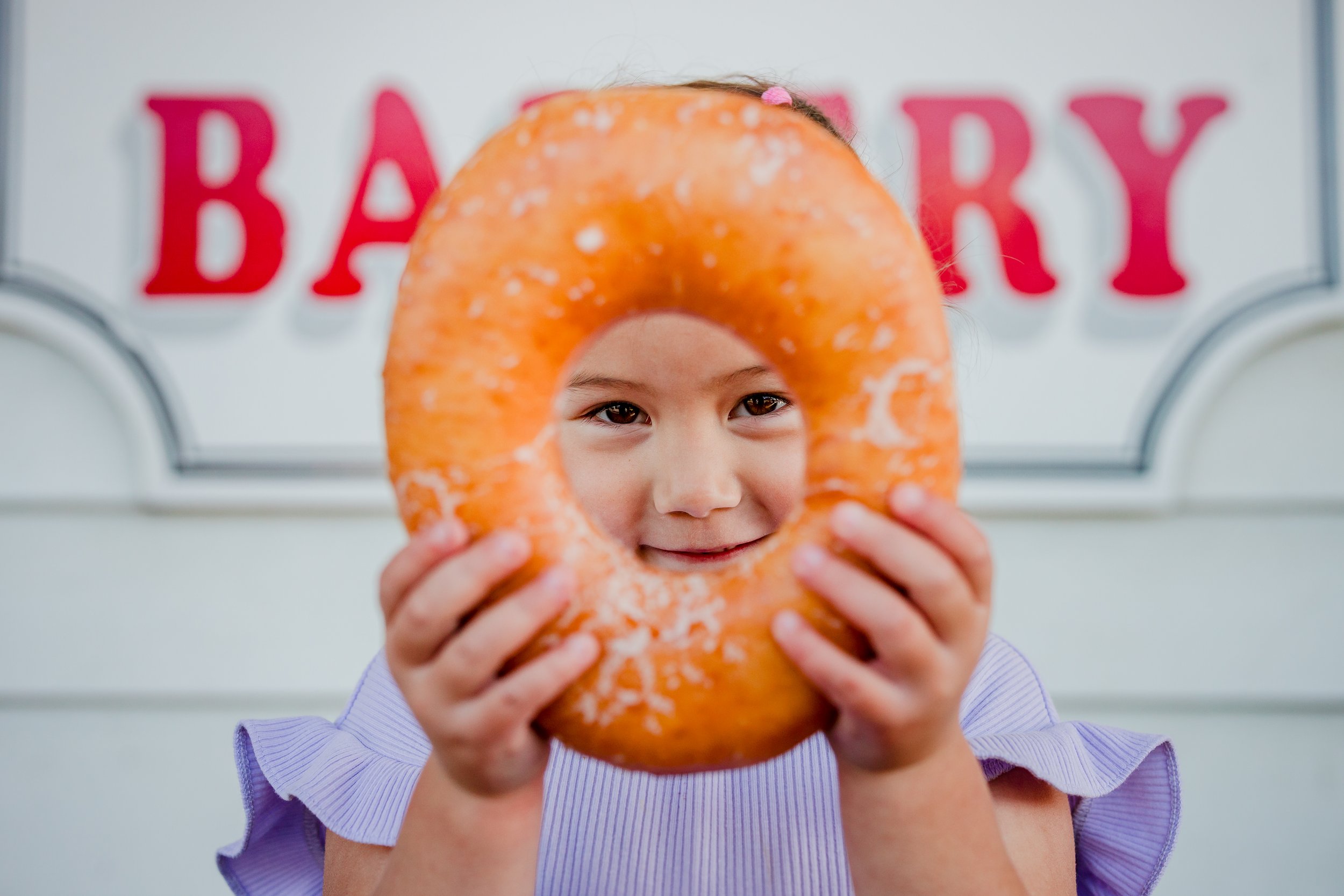 A young girl holds an orange donut with a hole in the center in front of her face, with a sign in the background that says 'Bakery' in red letters.