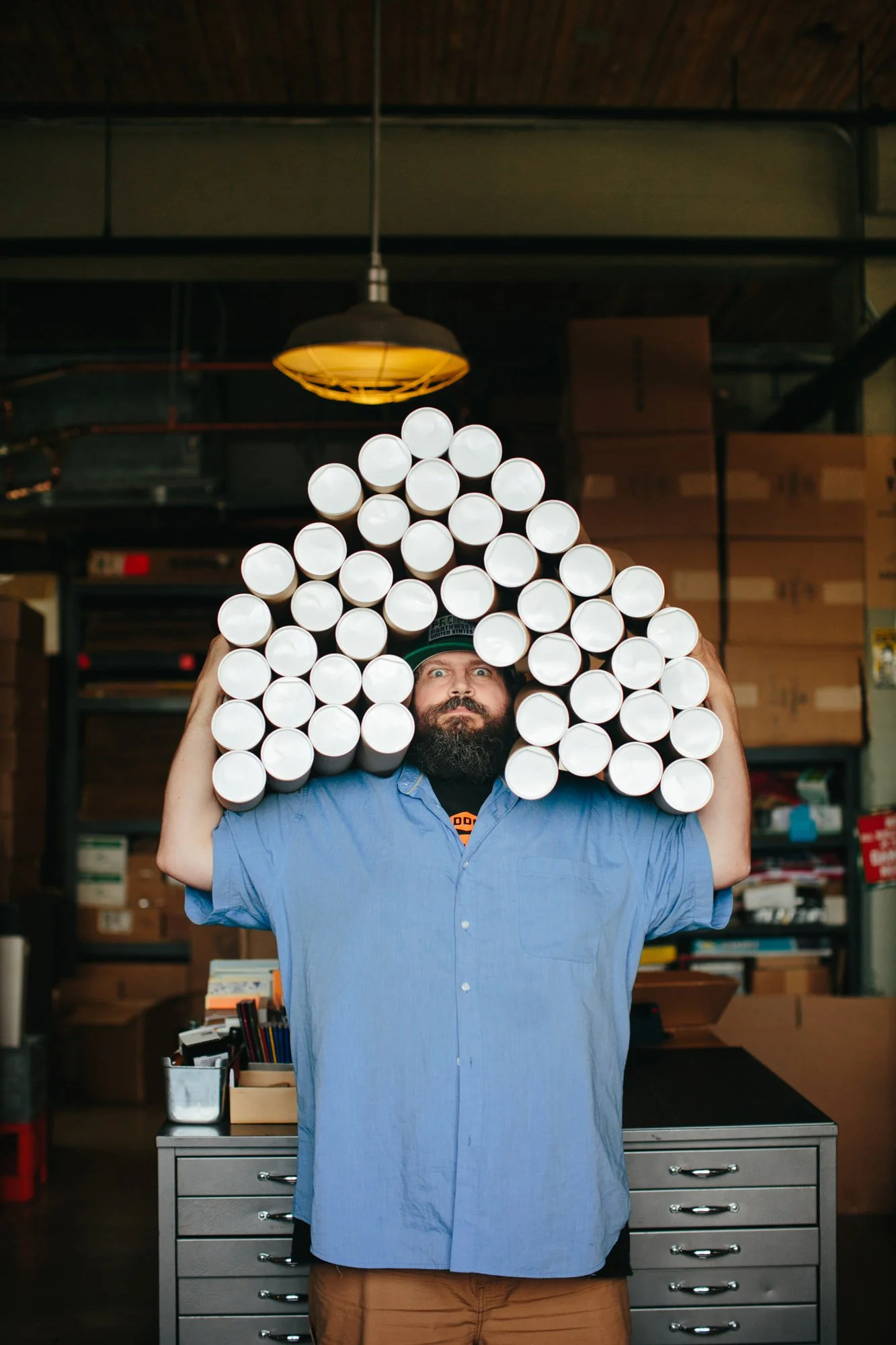 Man with a beard wearing a blue shirt carrying a large quantity of white cylindrical objects on his shoulders in a warehouse or storage room.