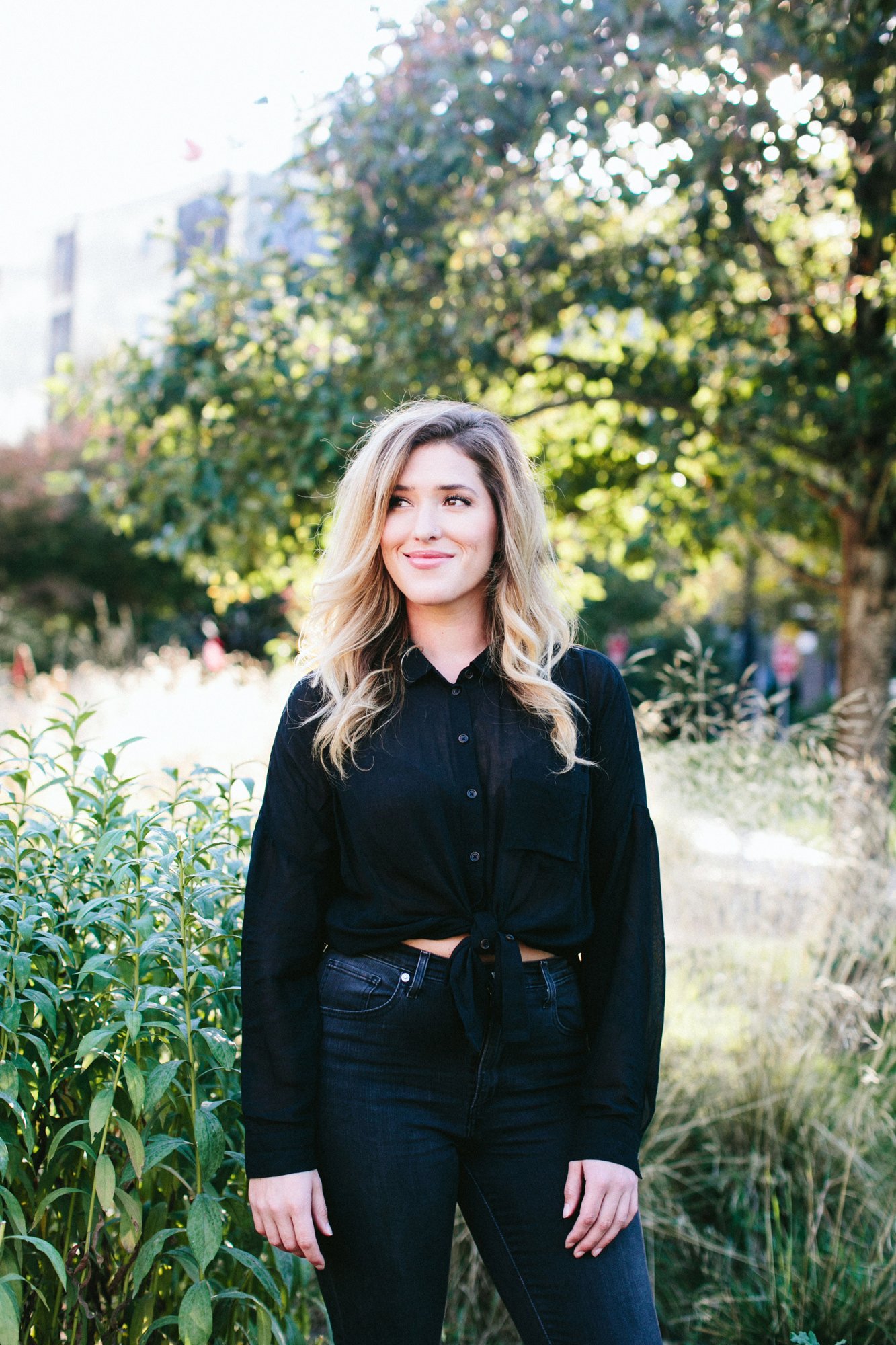 A young woman with blonde wavy hair smiling outdoors, wearing a black button-up shirt tied at the waist and dark jeans, surrounded by greenery and trees.
