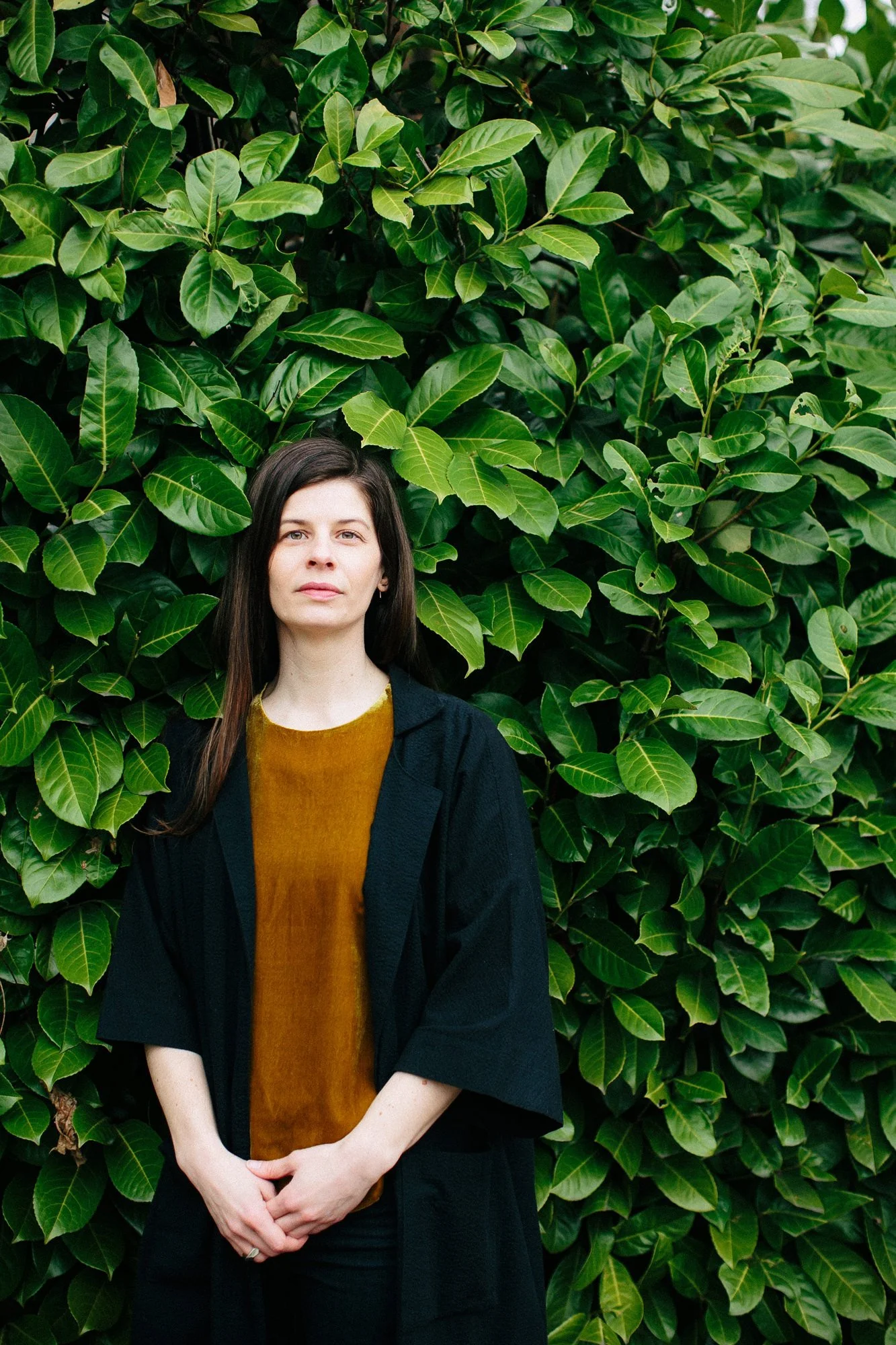 A woman with dark hair stands in front of a lush green leafy hedge, wearing a black coat over a mustard yellow top.