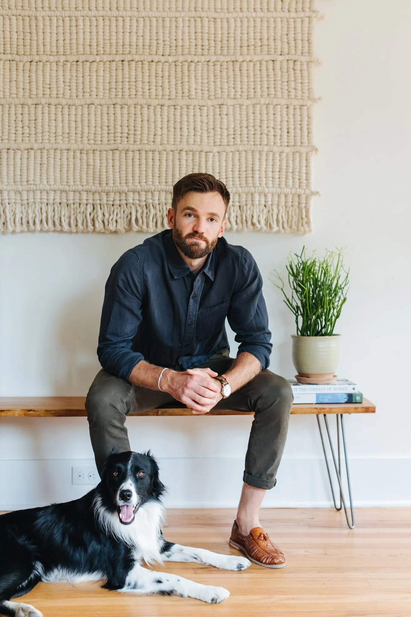 A man with a beard and wearing a dark shirt sitting on a wooden bench with a black and white dog lying on the floor in front of him. There is a potted plant on the bench and a woven wall hanging behind him.
