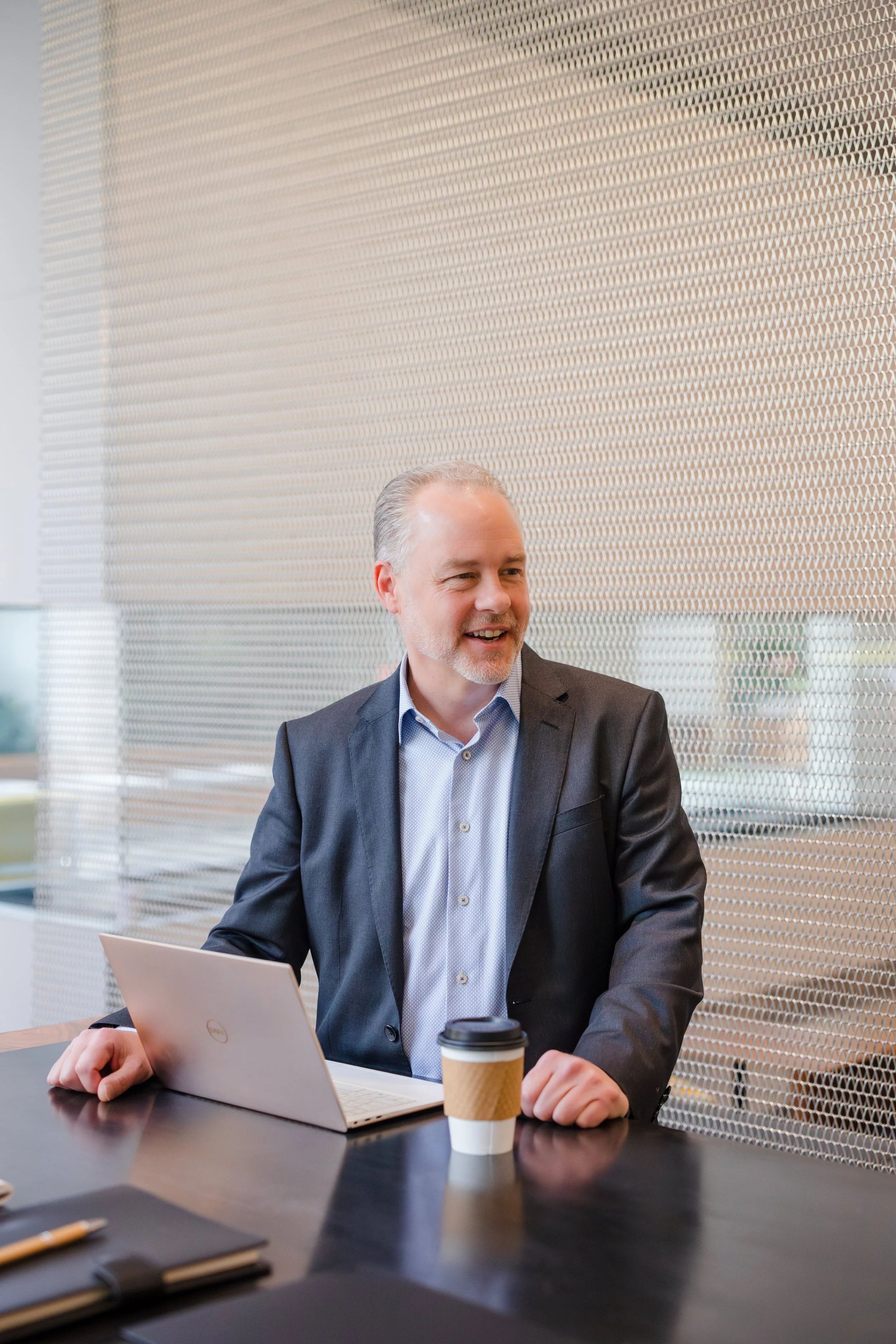 A middle-aged man with gray hair and a beard, dressed in a dark suit jacket and light blue shirt, sitting at a conference table with a laptop and a takeaway coffee cup, smiling and talking during a meeting in a modern office with a perforated metal w
