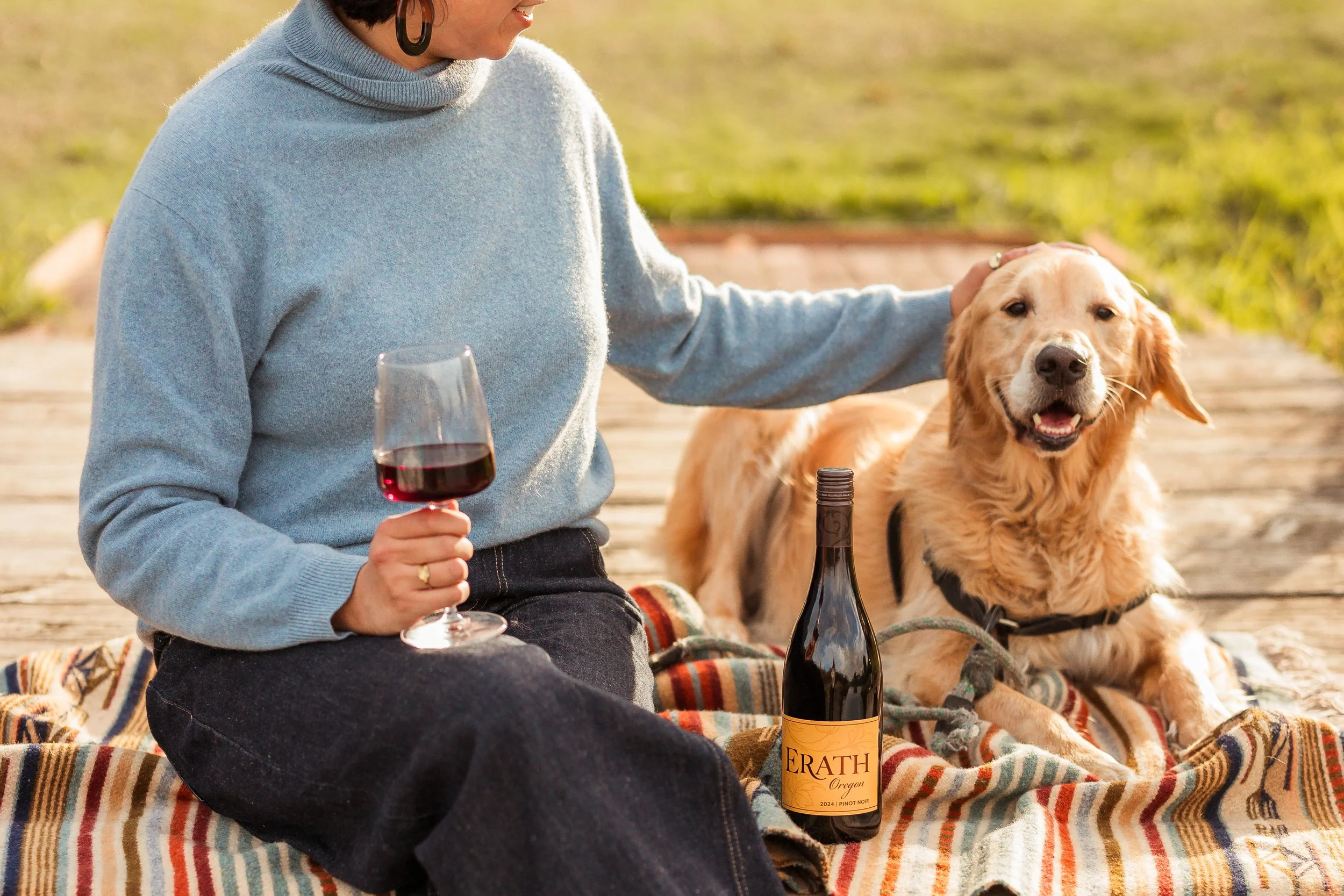 A woman holding a glass of red wine next to a golden retriever dog, sitting on a blanket with a bottle of wine on it, outdoors on a wooden surface.