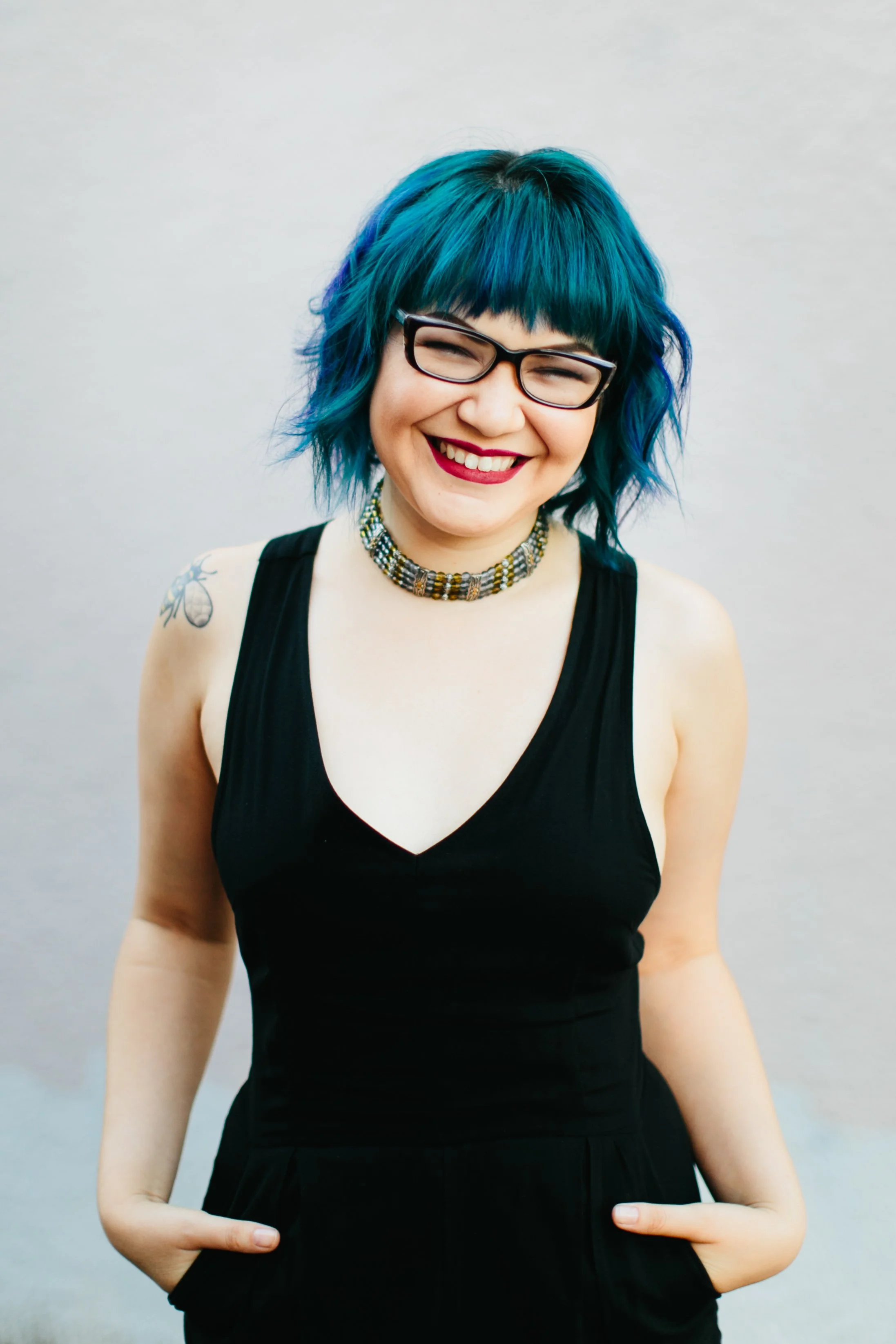 A woman with blue hair, glasses, and a black sleeveless dress, smiling with her eyes closed, standing against a plain light gray background.