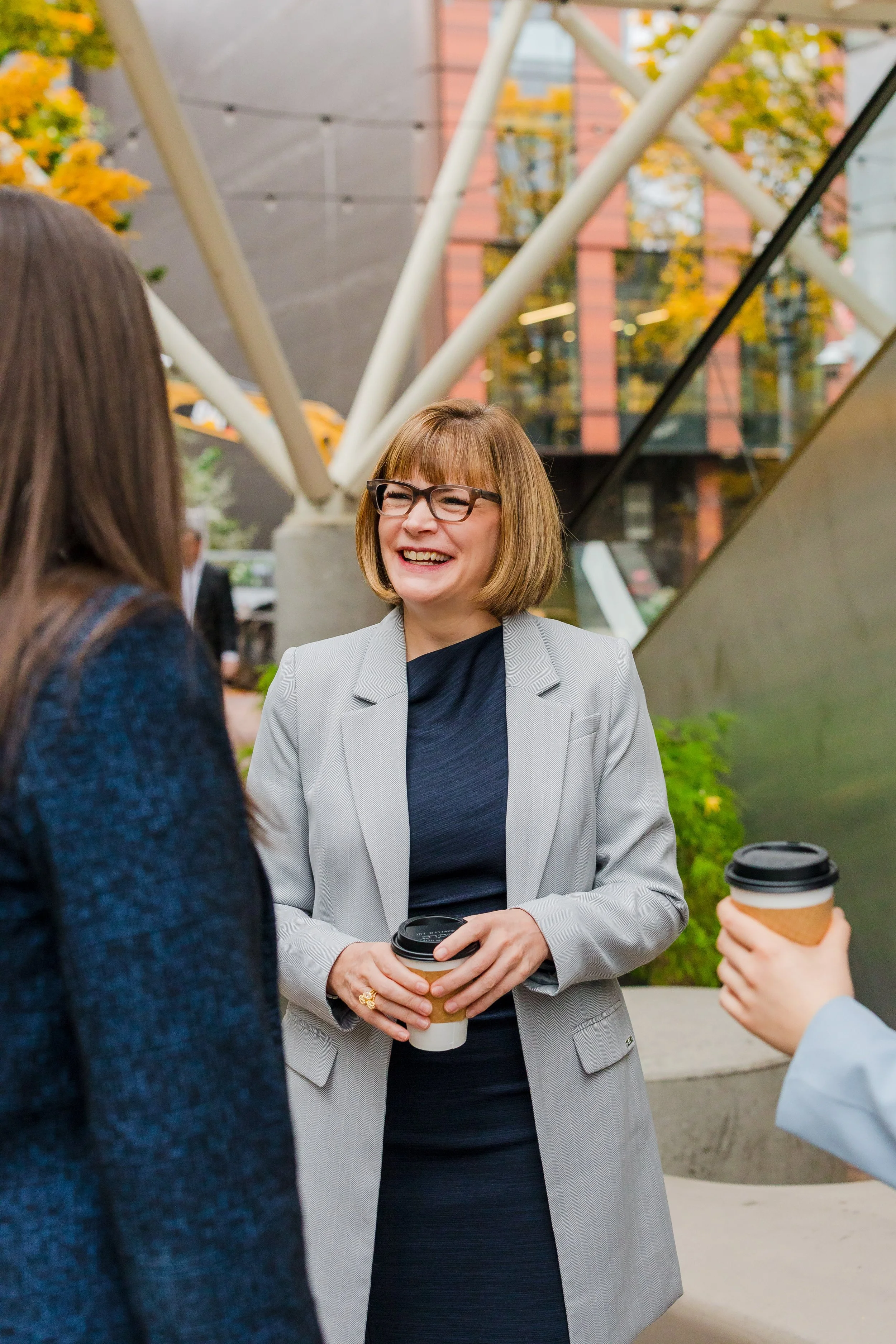 Three women holding coffee cups and talking outside near a modern building with large glass windows and structural beams.