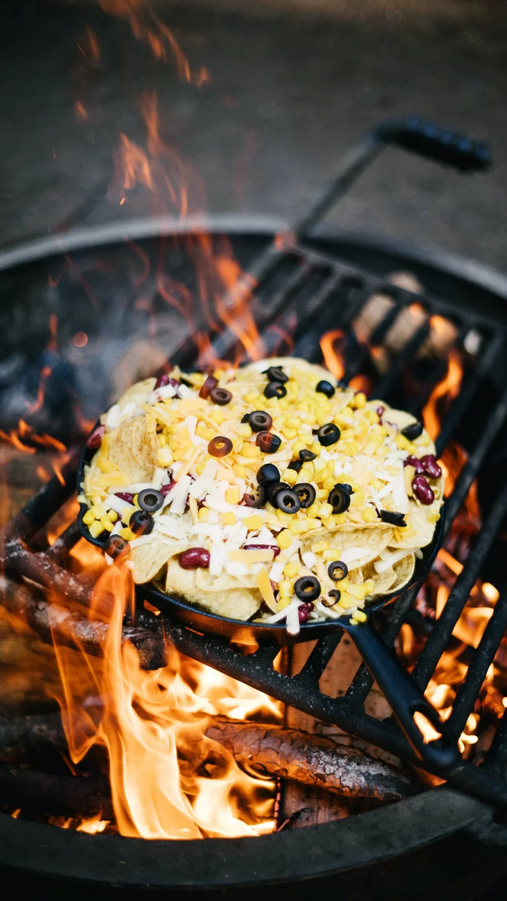 Tortilla chips topped with shredded cheese, black and red beans, and corn, being grilled over an open fire.