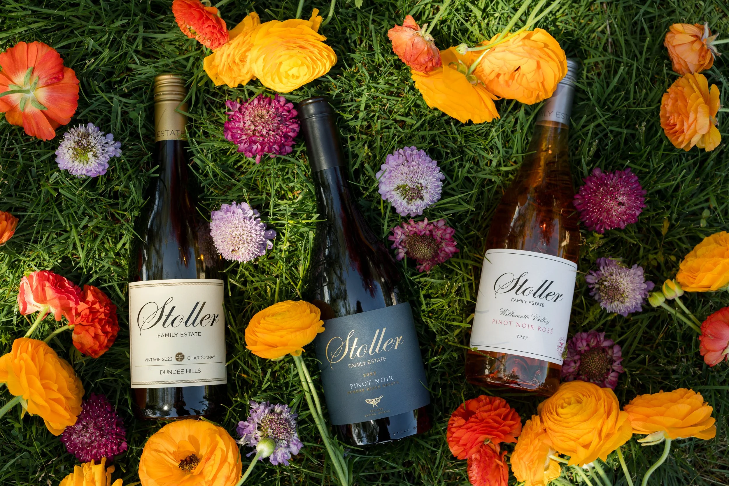 Three bottles of Stoller wine lying on green grass surrounded by orange, purple, and pink flowers.