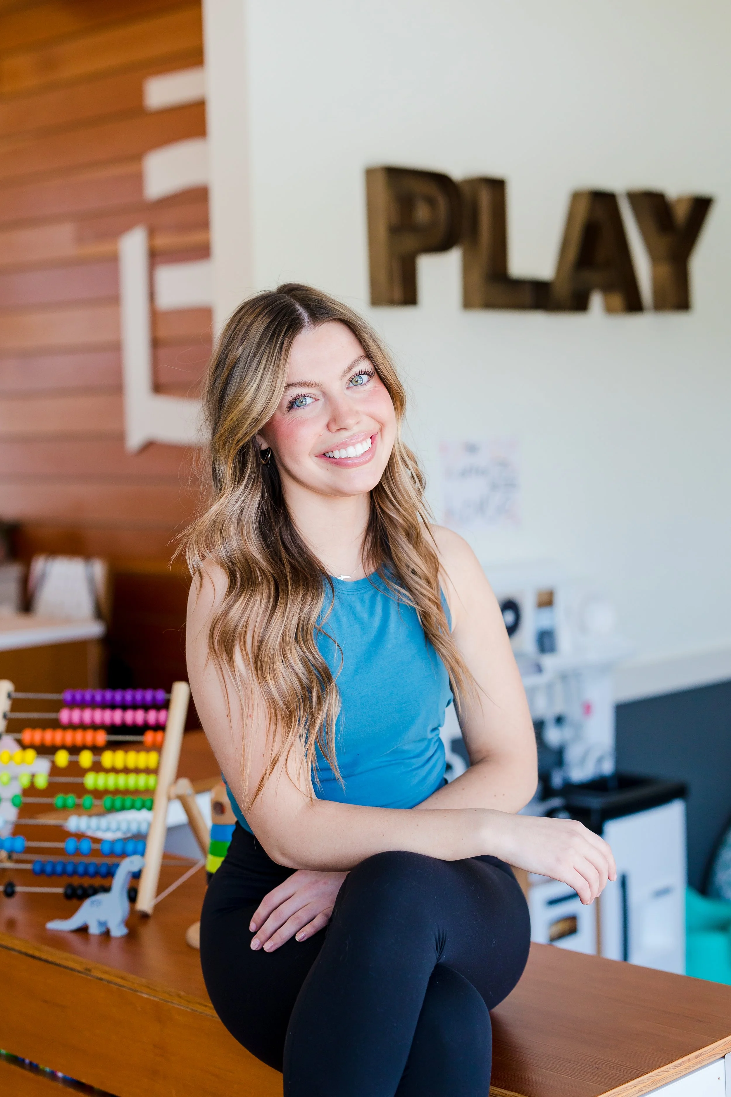 A young woman with long wavy blonde hair, wearing a blue sleeveless top and black leggings, sitting on a wooden counter with her legs crossed, smiling at the camera, in a play-themed room with colorful toys, abacus, and the word 'PLAY' on the wall.
