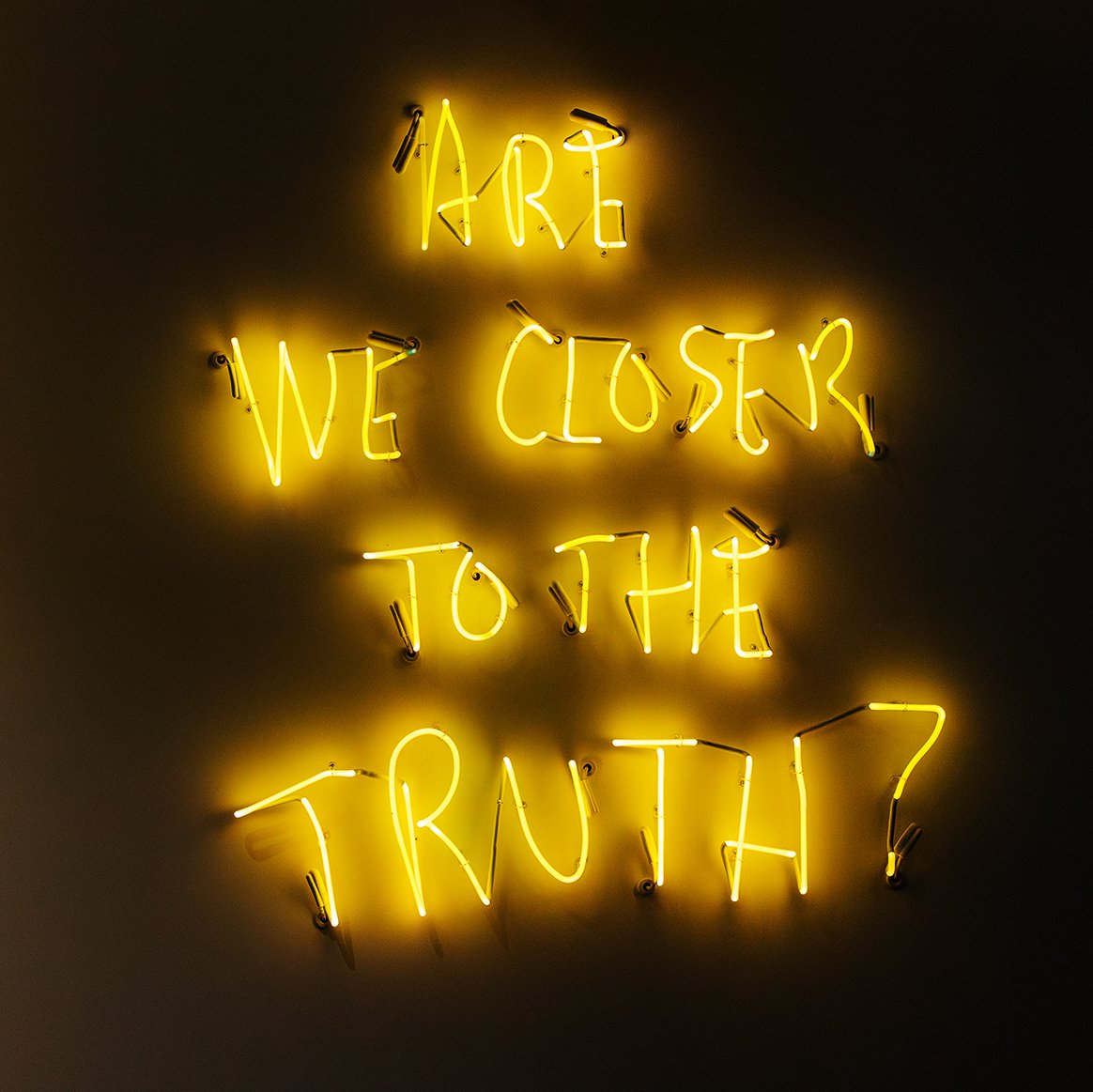 Neon sign reading 'Art, We Close To The Truth?' in yellow on a dark wall.