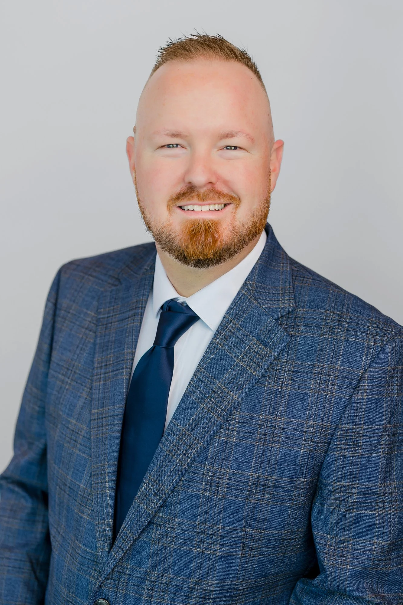 A man with a beard and short hair smiling, wearing a blue plaid suit, white shirt, and dark blue tie against a plain light grey background.