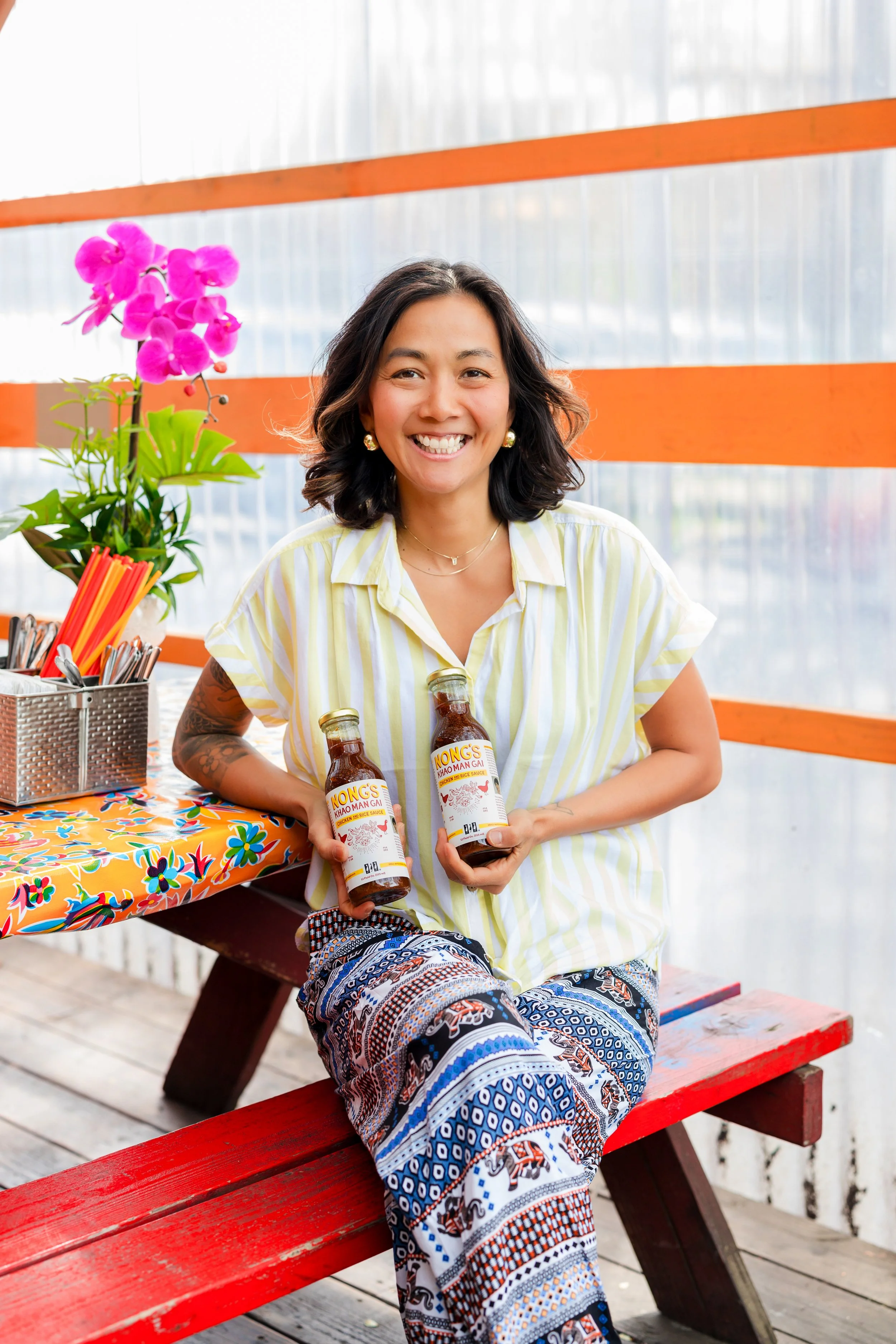A woman sitting on a red bench, smiling, holding two bottles of Nong's Khao Mām Gai, with a colorful table, a potted orchid, and utensils nearby in a bright, airy setting.