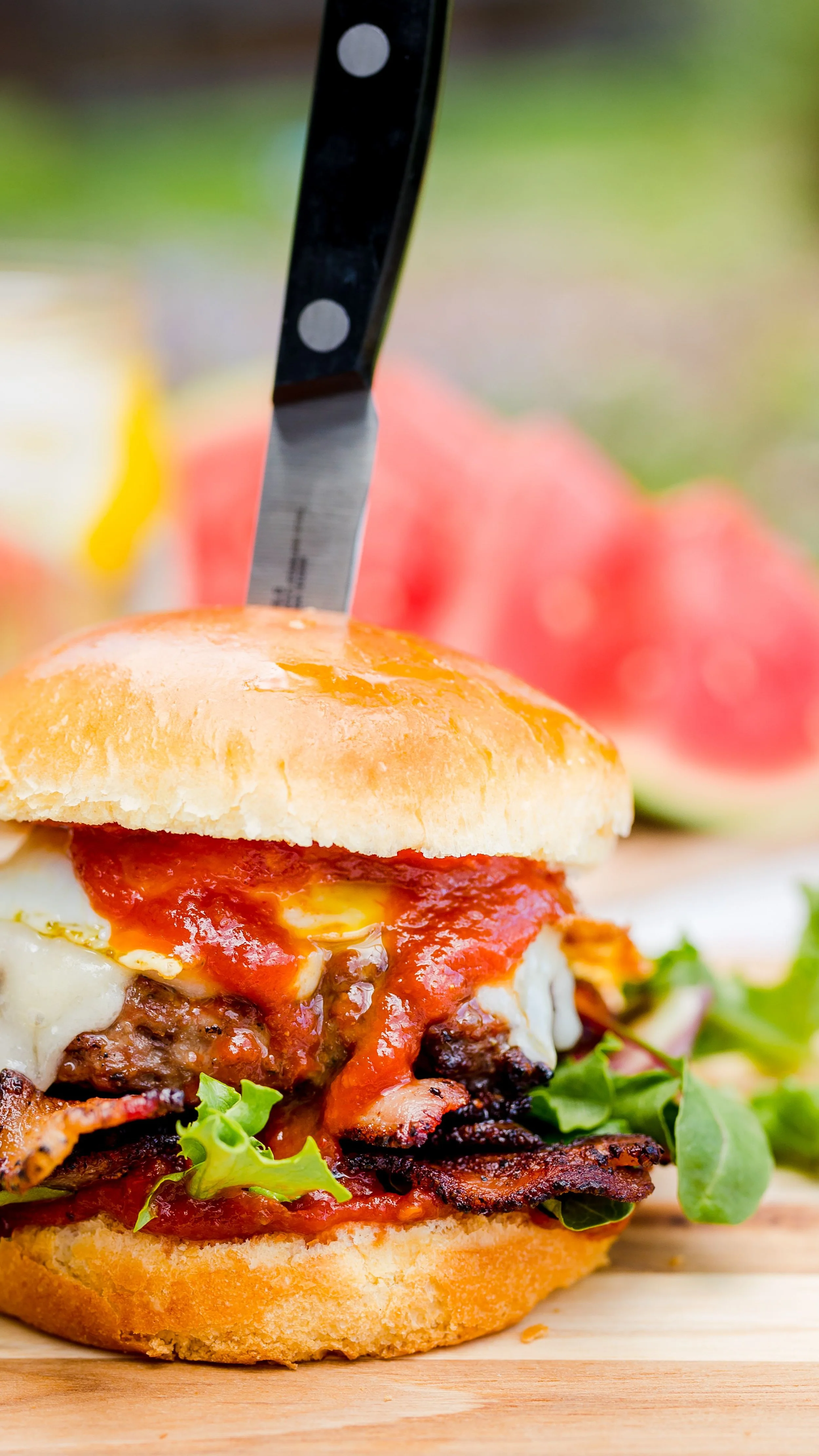 Close-up of a meatloaf sandwich with tomato sauce, bacon, cheese, and greens on a bun, with a knife stabbing through the top bun, on a wooden board with watermelon slices in the background.