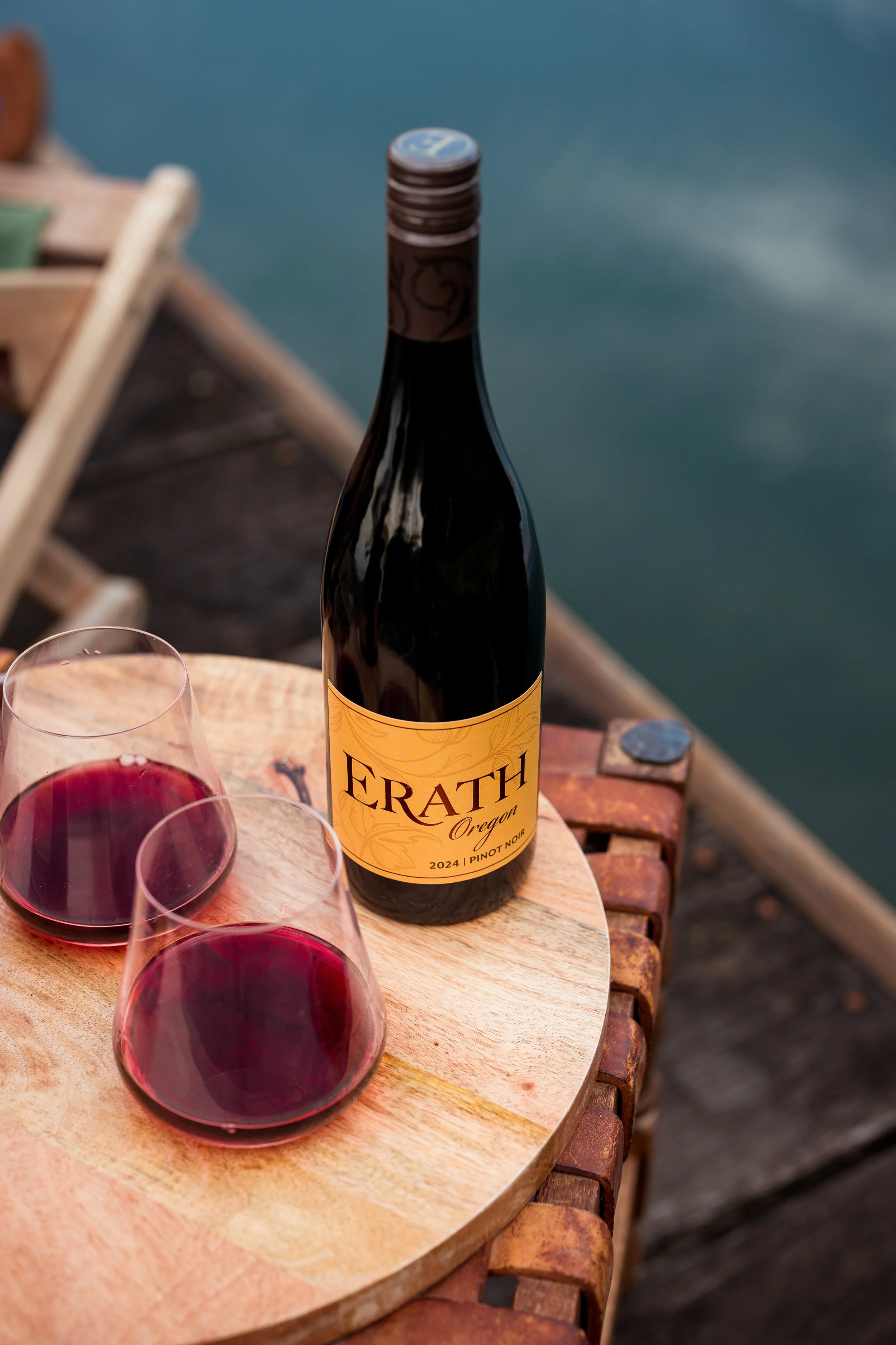 A bottle of Erath Oregon Pinot Noir 2024 and two glasses of red wine on a round wooden table near water.