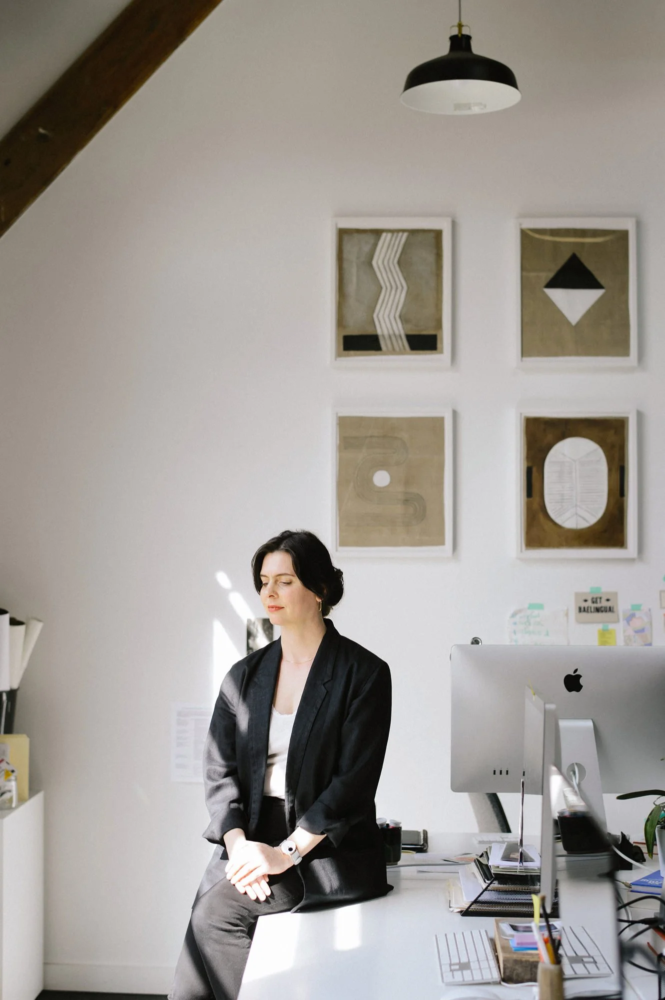 A woman sitting on the edge of a white desk in a modern office, with sunlight casting shadows. She has dark hair, wears a black blazer and pants, and is looking down with eyes closed.