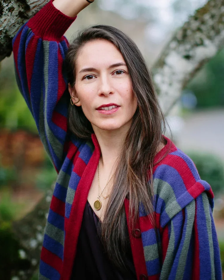 A woman with long brown hair wearing a colorful striped sweater over a black top, standing outdoors, with trees in the background.
